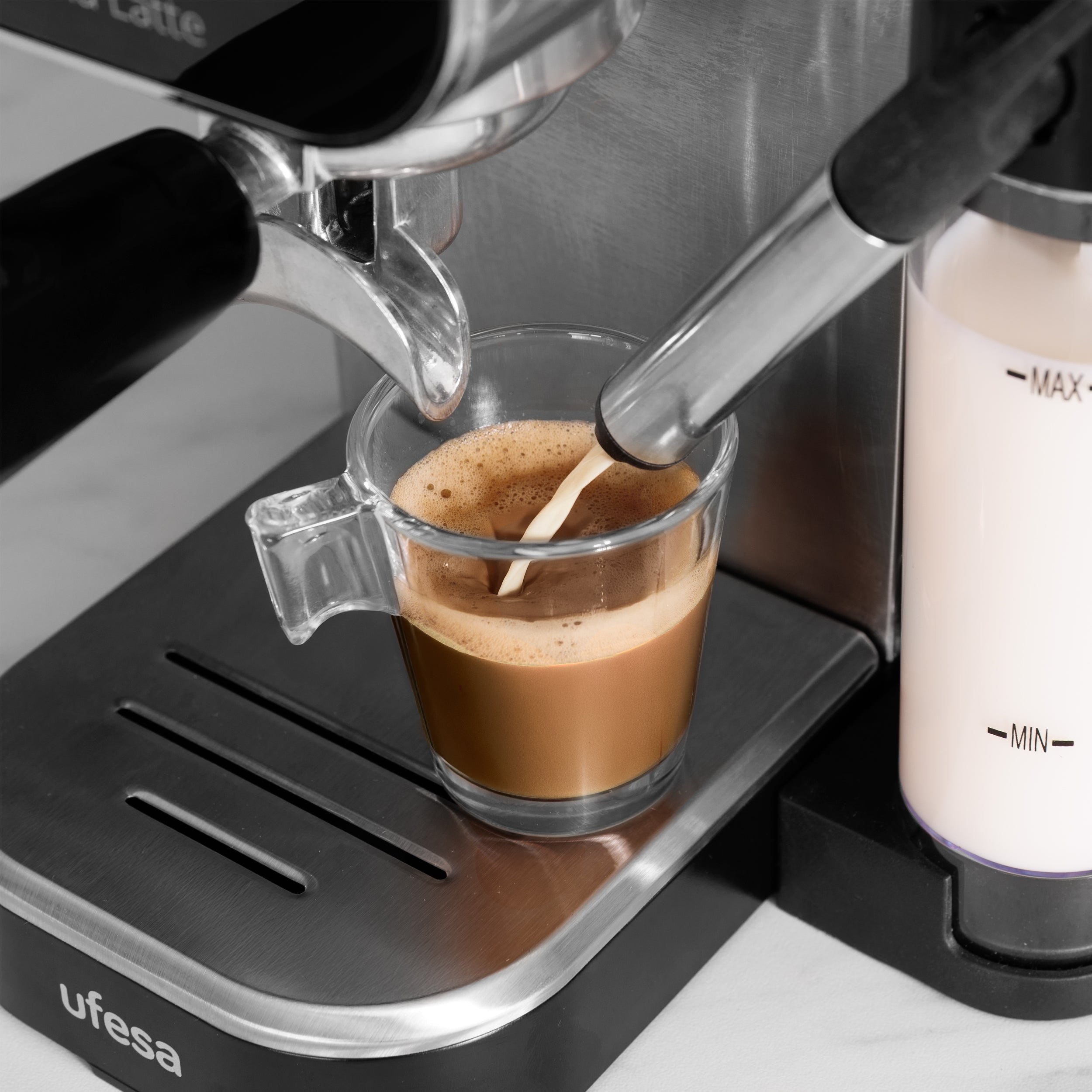 Una máquina de café vierte leche en una taza de café de vidrio. Se ve espuma en la parte superior. La máquina es plateada y negra.