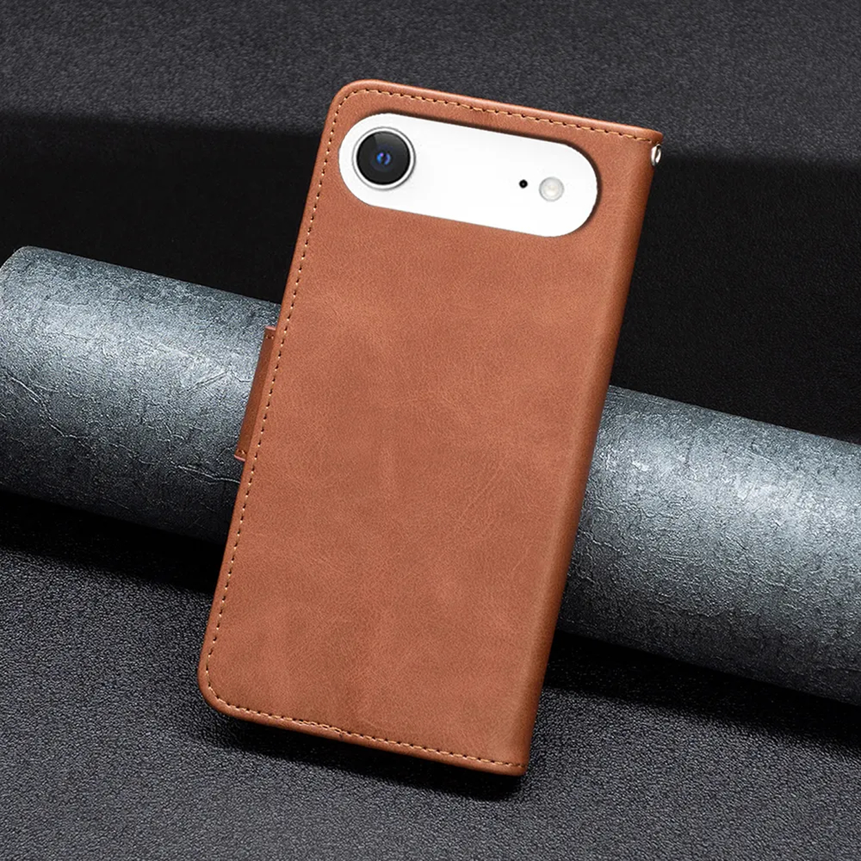 Een bruine leren telefoonhoes met een witte camerasectie, liggend op een donker oppervlak met een grijs cilindrisch object.