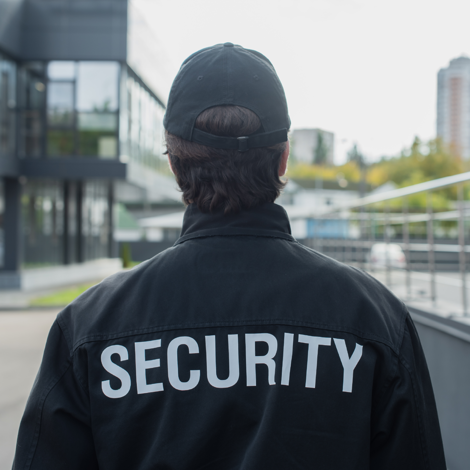 Un agent de sécurité portant un uniforme et une casquette noirs, avec le mot 'SECURITY' dans le dos. Bâtiment et arbres en arrière-plan.