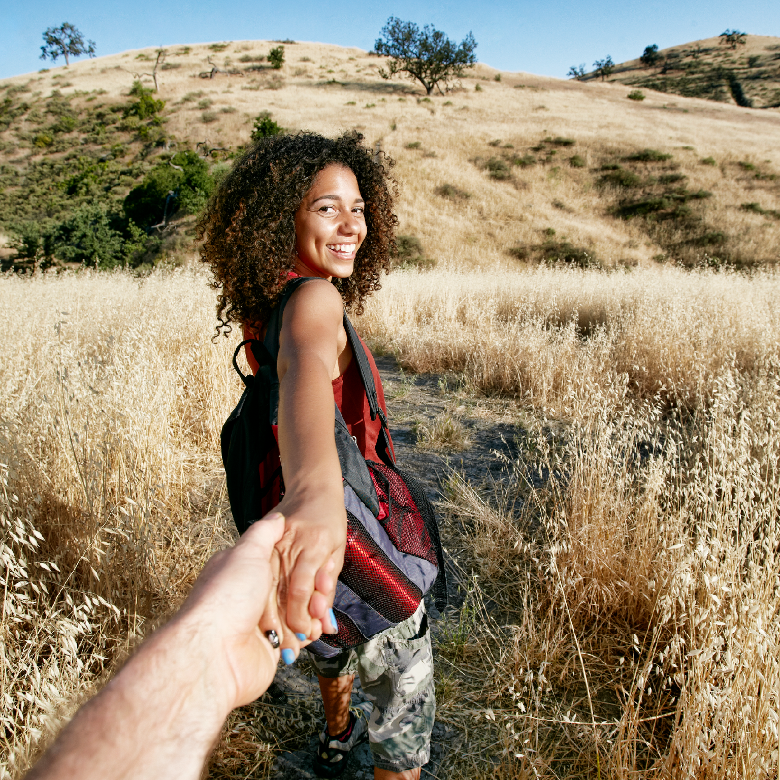 A woman with curly hair smiles while holding a hand. They are on a trail in a field of dry grass, with a hill in the background.