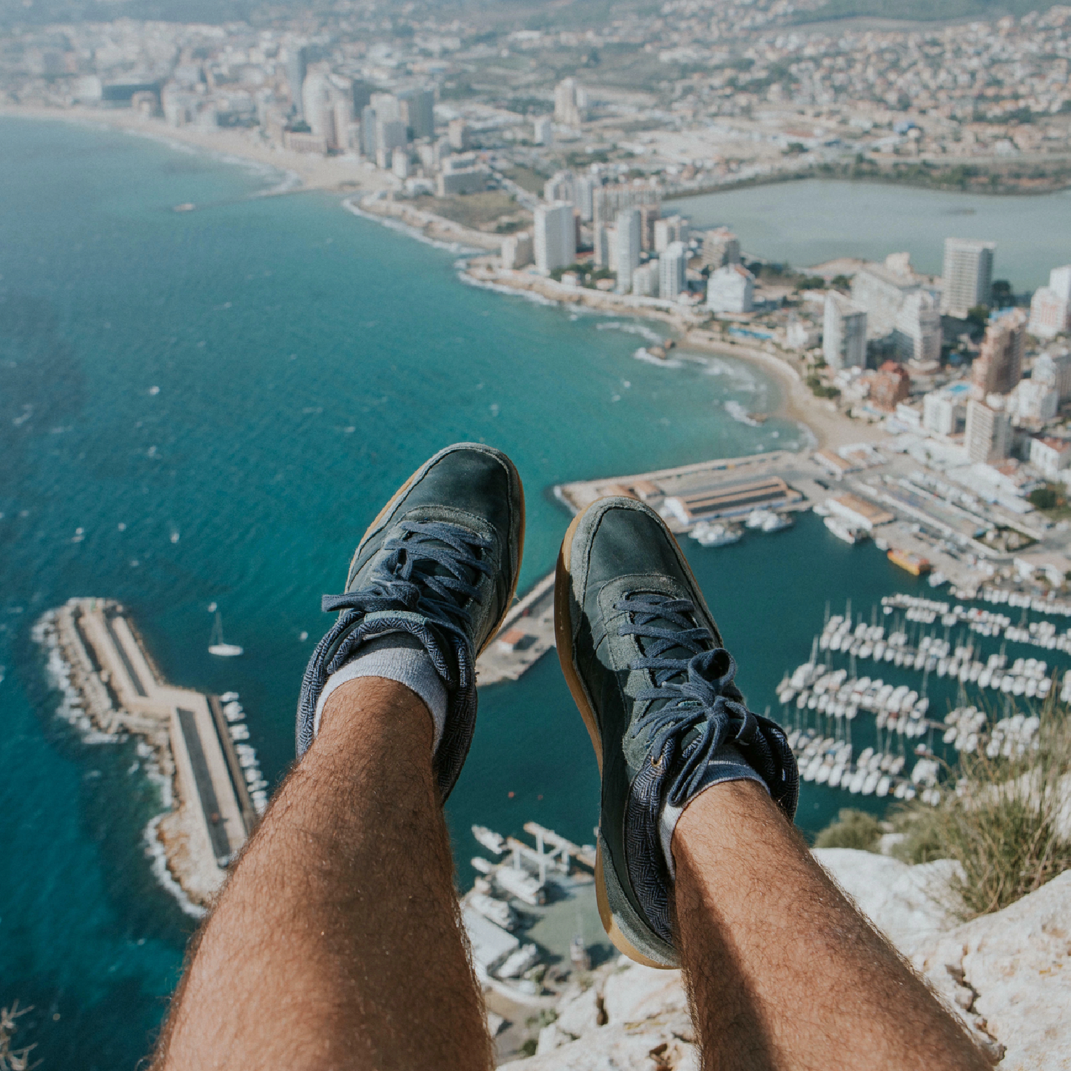 Feet in blue shoes over a coastal city. Buildings, sea, and boats visible. Hairy legs and a cliff are in the foreground.