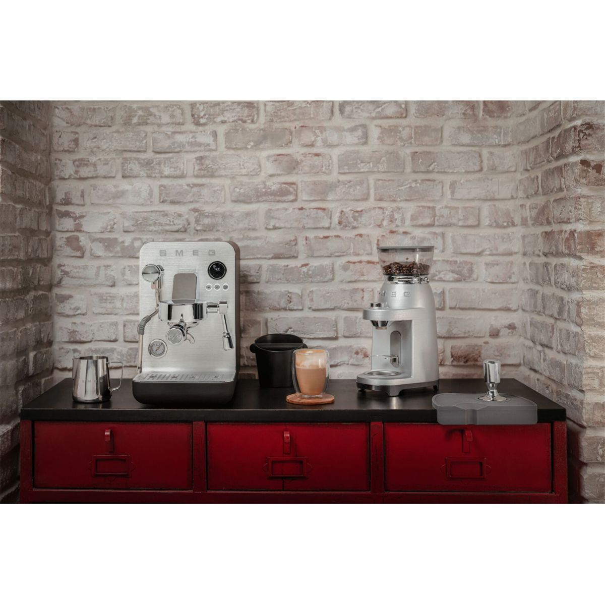 Srebrny ekspres do kawy i młynek do kawy na czerwonej szafce na tle ceglanej ściany. Obok młynka stoi filiżanka kawy.