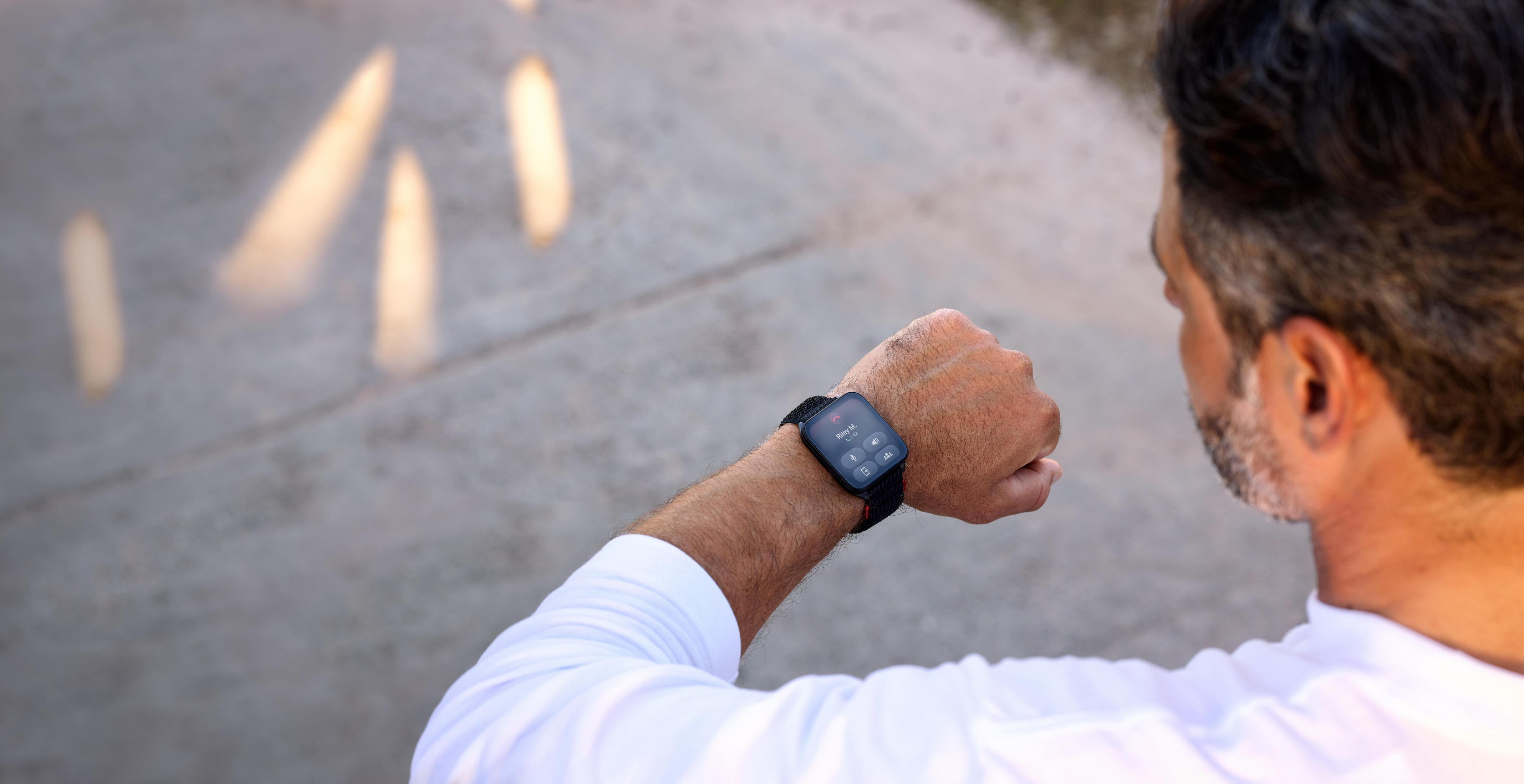 Ein Mann, der eine Smartwatch trägt, schaut darauf. Die Uhr zeigt einen Anruf von Riley M. Der Hintergrund ist verschwommener Beton.