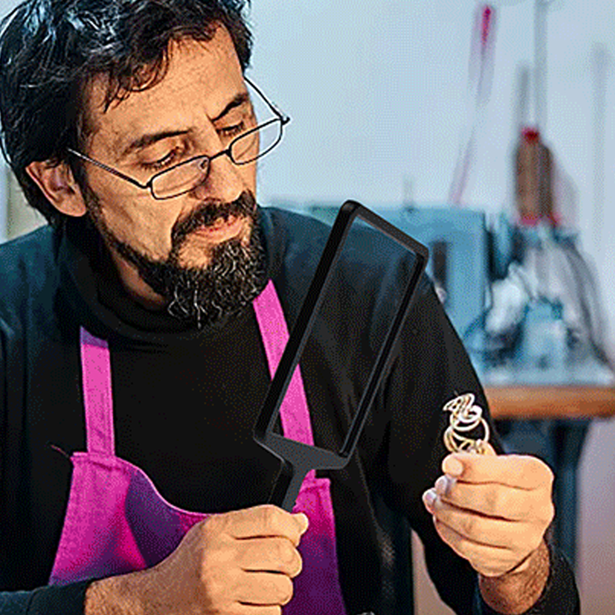 Ein Mann mit Brille betrachtet Schmuck durch eine Lupe. Er trägt ein schwarzes Hemd und eine rosa Schürze. Der Hintergrund ist eine Werkstatt.