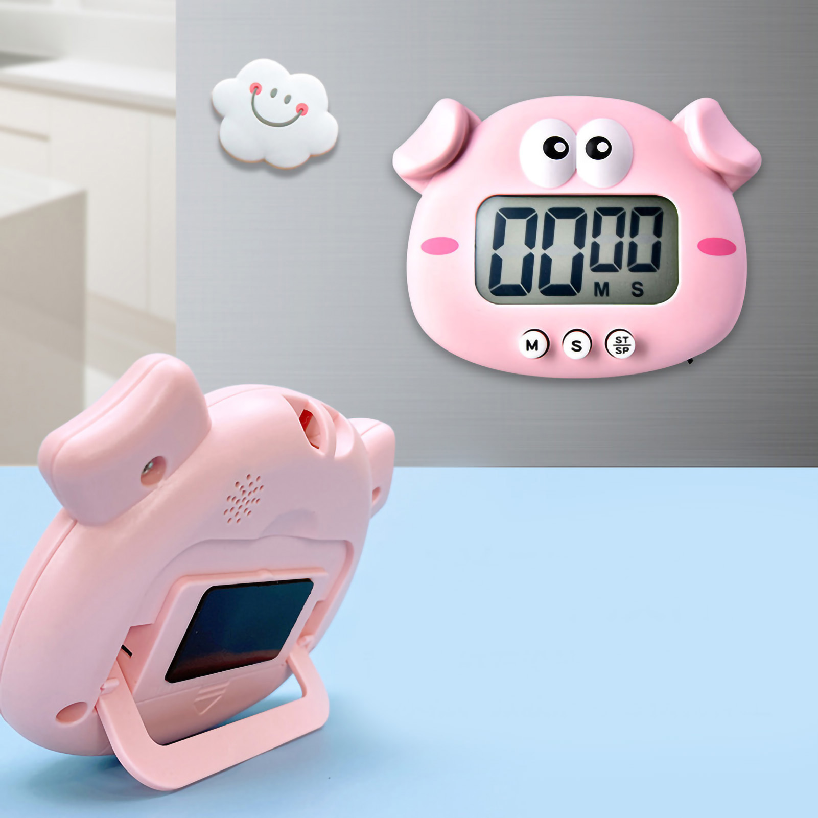 Ein rosa, schweinähnlicher Küchentimer zeigt 00:00 an. Ein lächelnder Wolkenmagnet ist darüber, ein zweiter Timer liegt auf blauem Untergrund.
