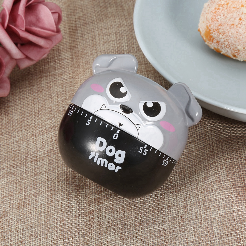 Ein hundeformiger Küchenwecker mit schwarz-grauem Design. Er liegt auf einer braunen, strukturierten Oberfläche neben einer rosa Blume und einem Teller mit Essen.