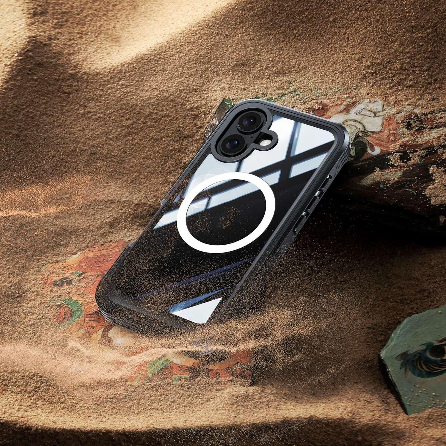 Eine schwarze Handyhülle mit weißem Ring und Kameralinsen liegt auf Sand. Der Hintergrund hat braune und grüne Elemente.