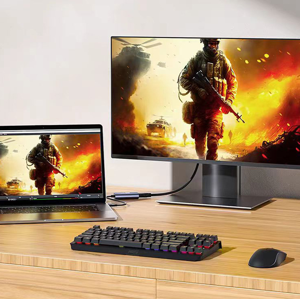 Ein Laptop und ein Monitor zeigen einen Soldaten in einer Kriegsszene. Auf einem Holzschreibtisch befinden sich eine Tastatur und eine Maus.