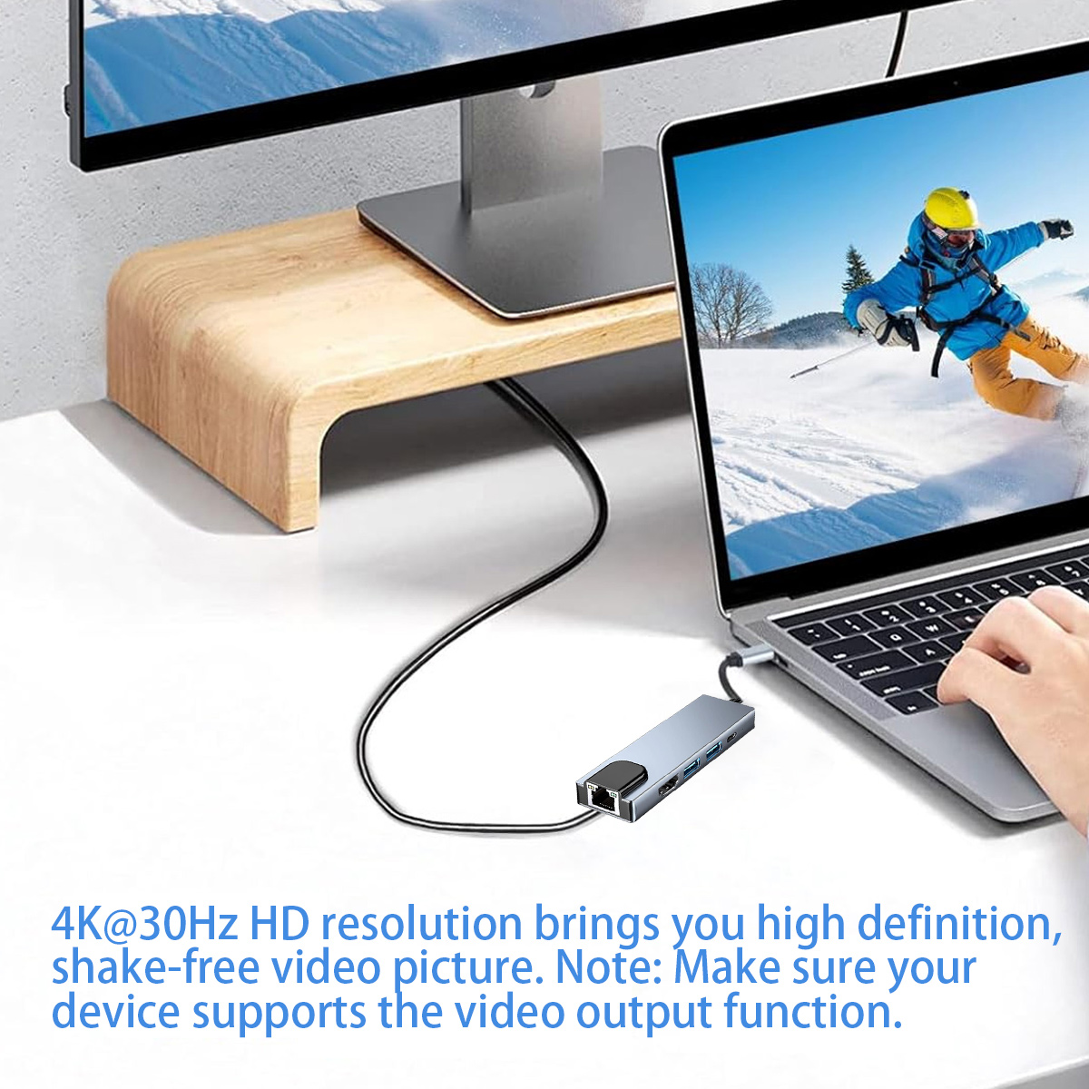 Laptop mit Hub verbunden, zeigt einen Skifahrer auf dem Bildschirm. Ein Monitor und Holzständer im Hintergrund.