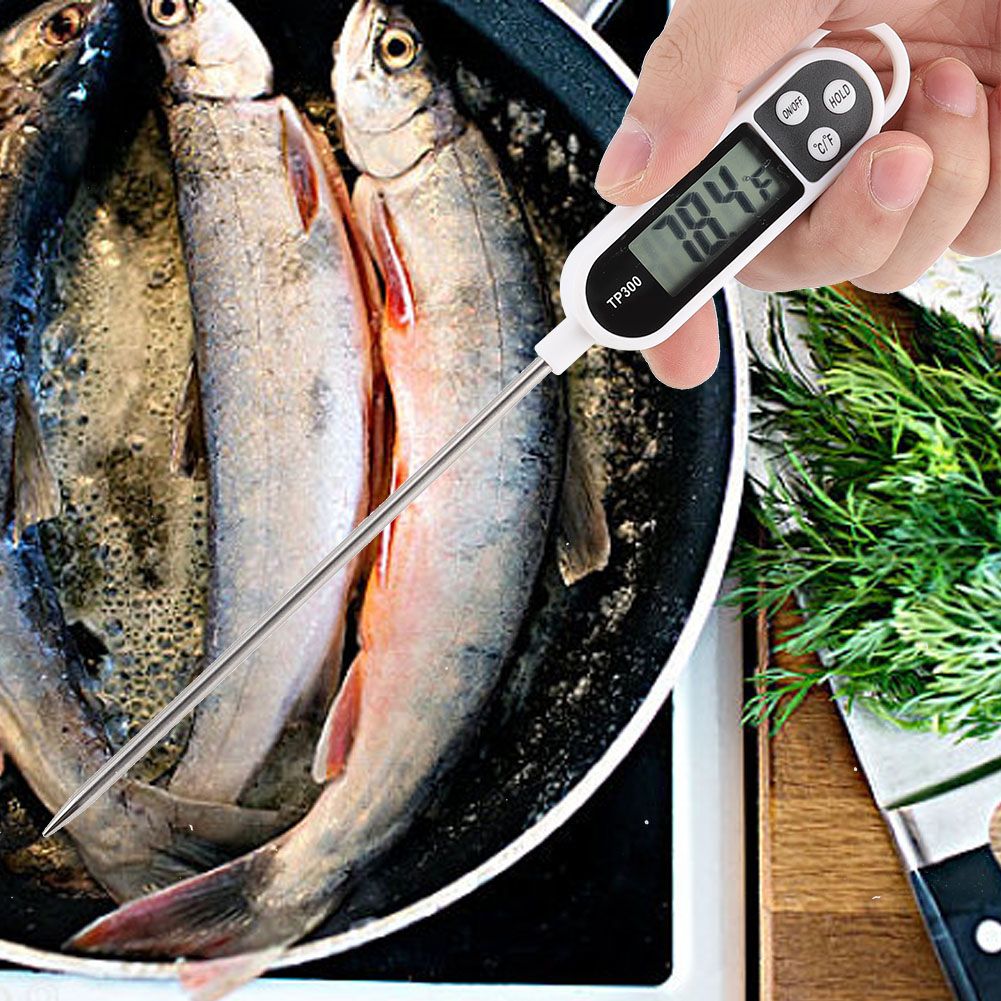 Eine Hand hält ein Lebensmitteltemperaturmessgerät, das in Fisch in einer Pfanne steckt. Das Thermometer zeigt 78,4 an. Grünes Kraut und ein Messer sind in der Nähe.