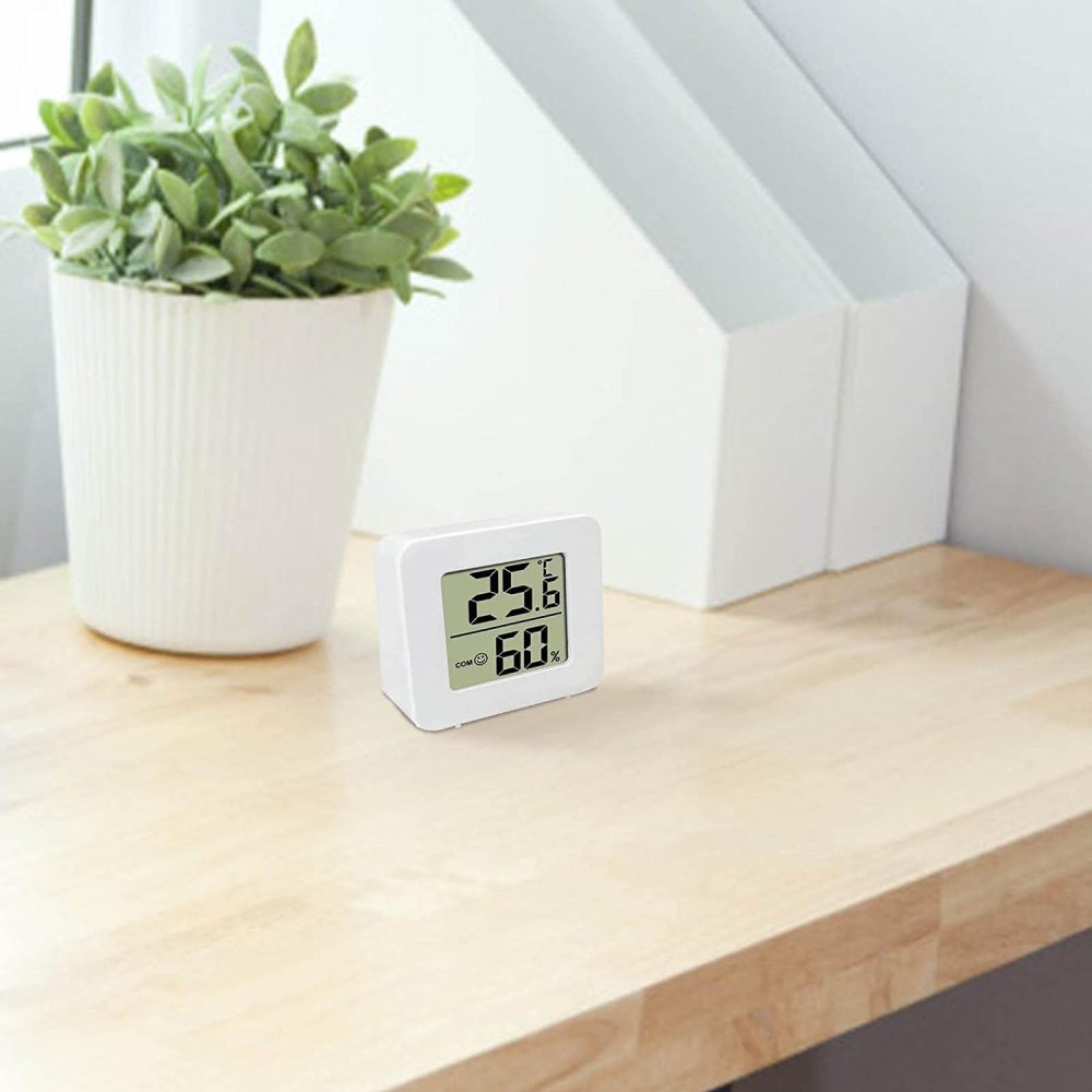 Ein weißes digitales Thermometer zeigt 25,6°C und 60 % Luftfeuchtigkeit an. Es steht auf einer Holzoberfläche in der Nähe einer Topfpflanze und weißer Aufbewahrung.