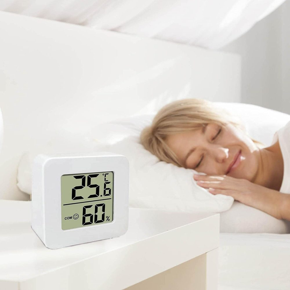 Eine Frau schläft im Bett neben einem weißen digitalen Thermometer. Das Thermometer zeigt Temperatur und Luftfeuchtigkeit an. Die Szene ist in einem Schlafzimmer.