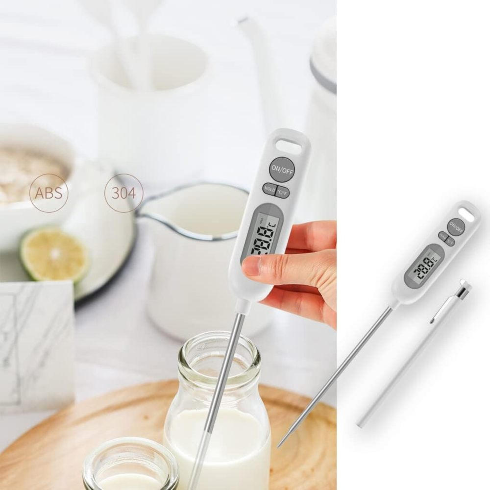 Eine Hand hält ein digitales Thermometer in Milch. Der Bildschirm zeigt 28,8 °C an. Weitere Objekte sind ein Glas Milch, Zitrone und weißes Geschirr.