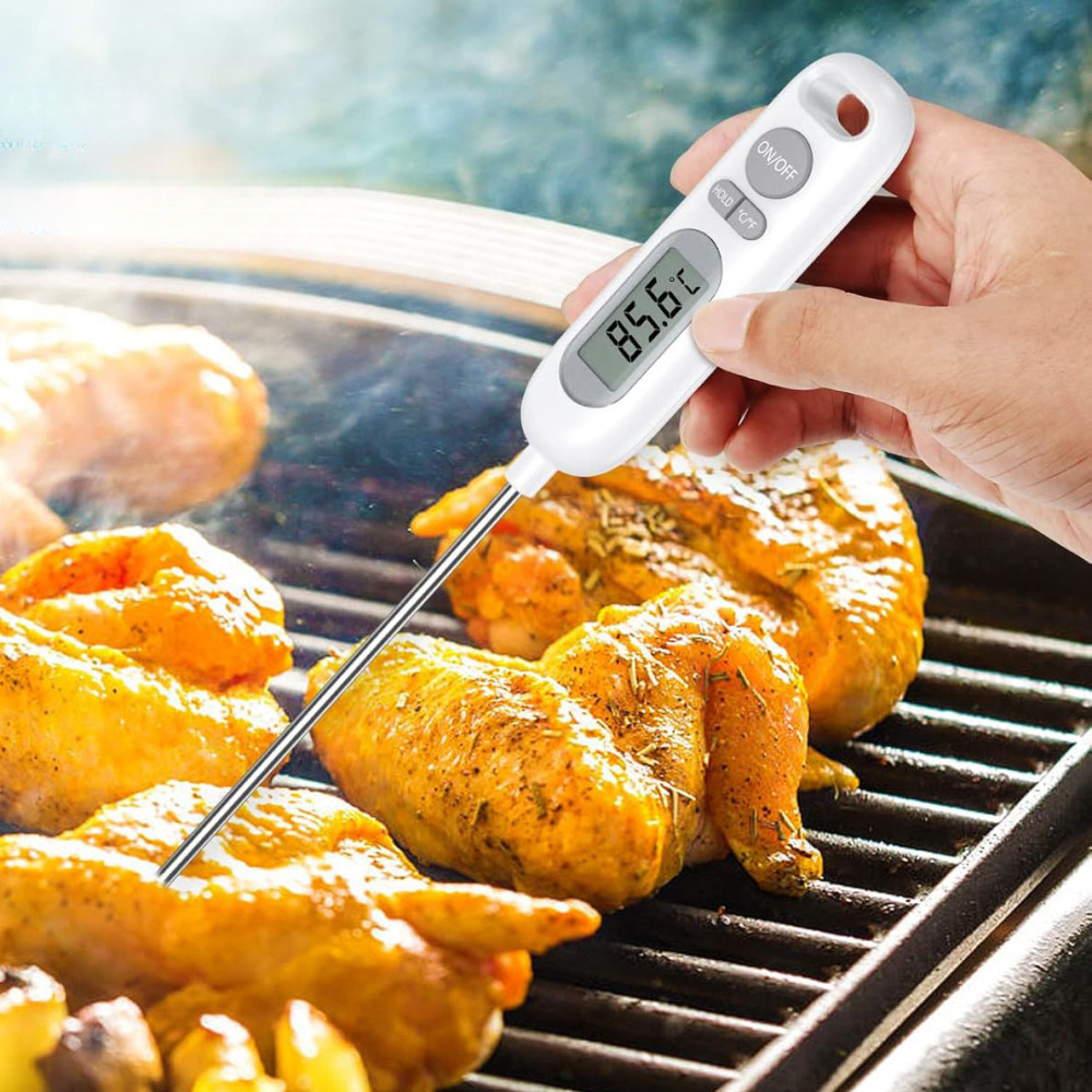 Eine Hand hält ein weißes Thermometer, das die Temperatur von gegrilltem Hähnchen auf einem Grill misst. Das Thermometer zeigt 85,6°C an.