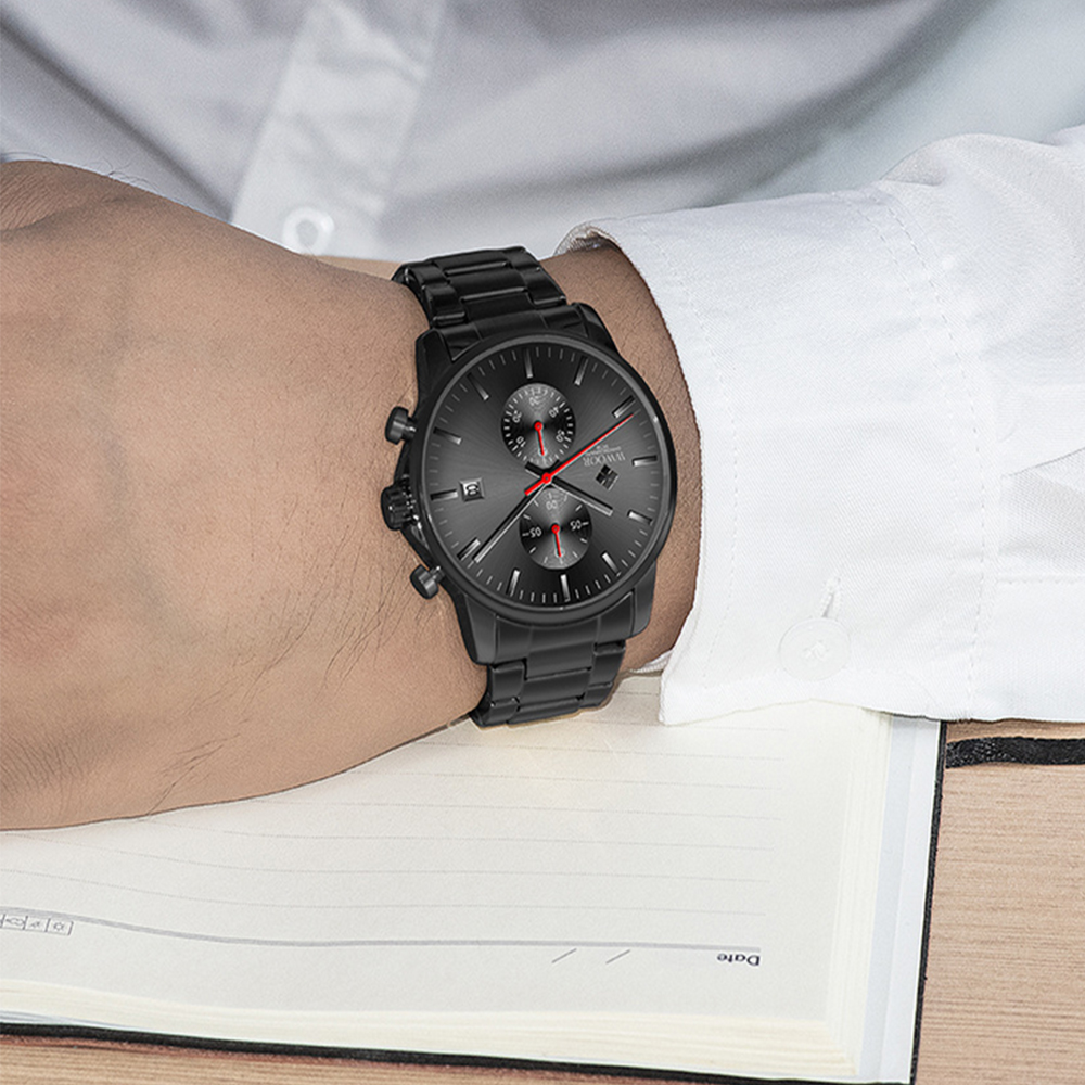 Eine schwarze Armbanduhr mit roten Zeigern befindet sich am Handgelenk eines Mannes. Er trägt ein weißes Hemd. Darunter liegt ein Notizbuch.