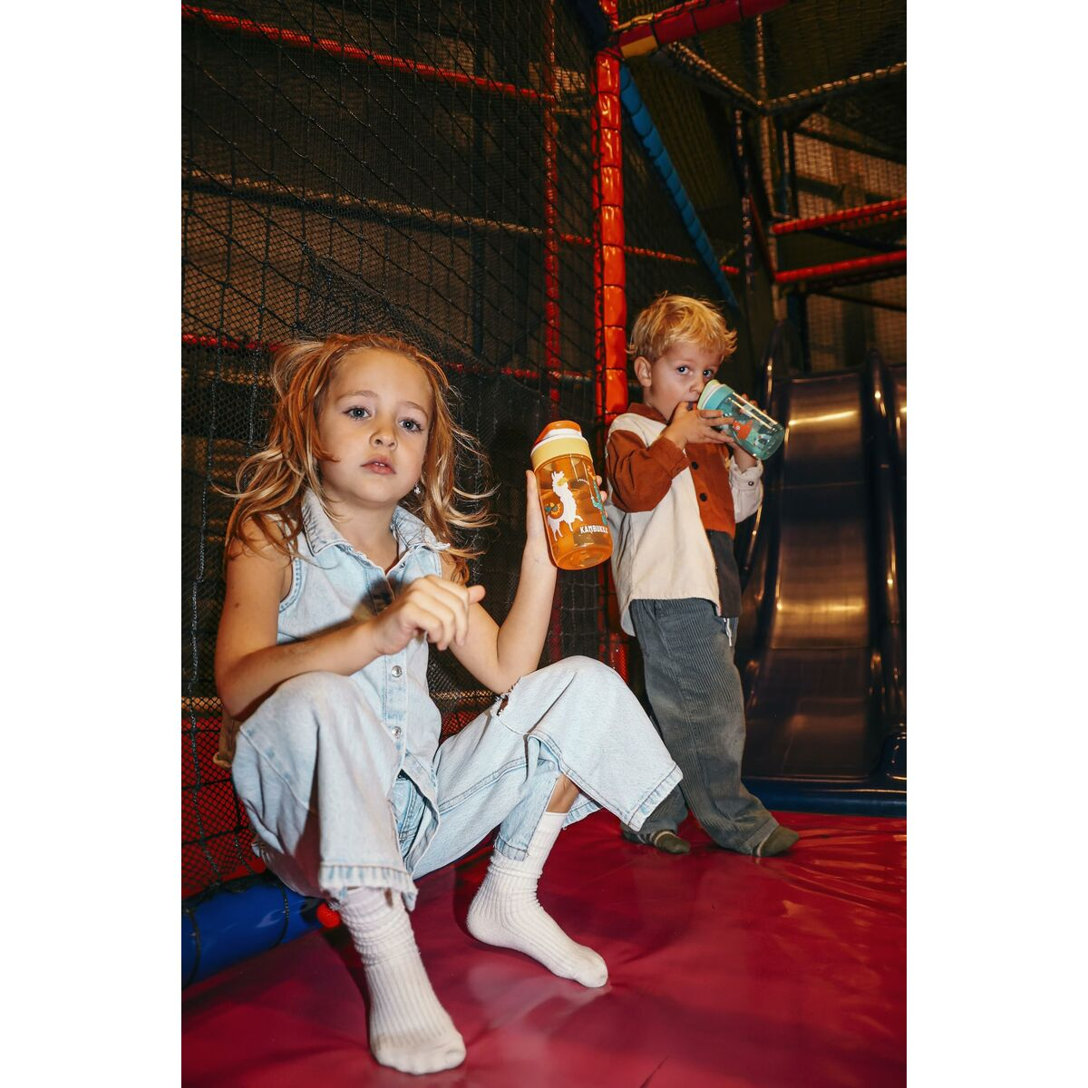 Zwei Kinder auf einem Spielplatz. Ein Mädchen sitzt und hält eine Wasserflasche. Ein Junge steht an einer Rutsche und trinkt.