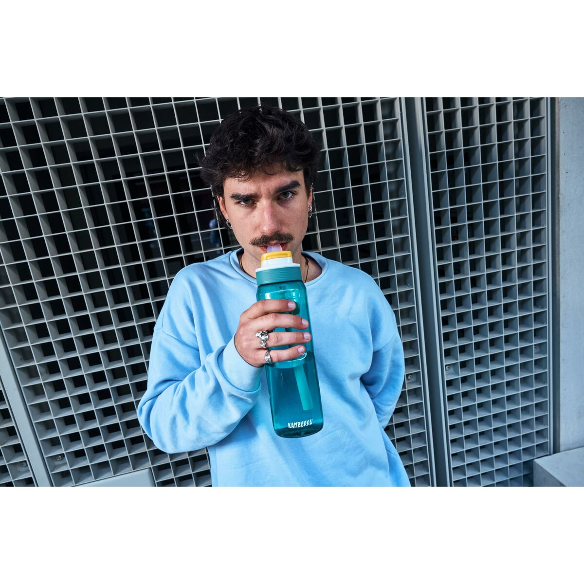 Ein Mann trinkt aus einer blauen Wasserflasche. Er trägt einen hellblauen Pullover und steht vor einem grauen Gitterhintergrund.