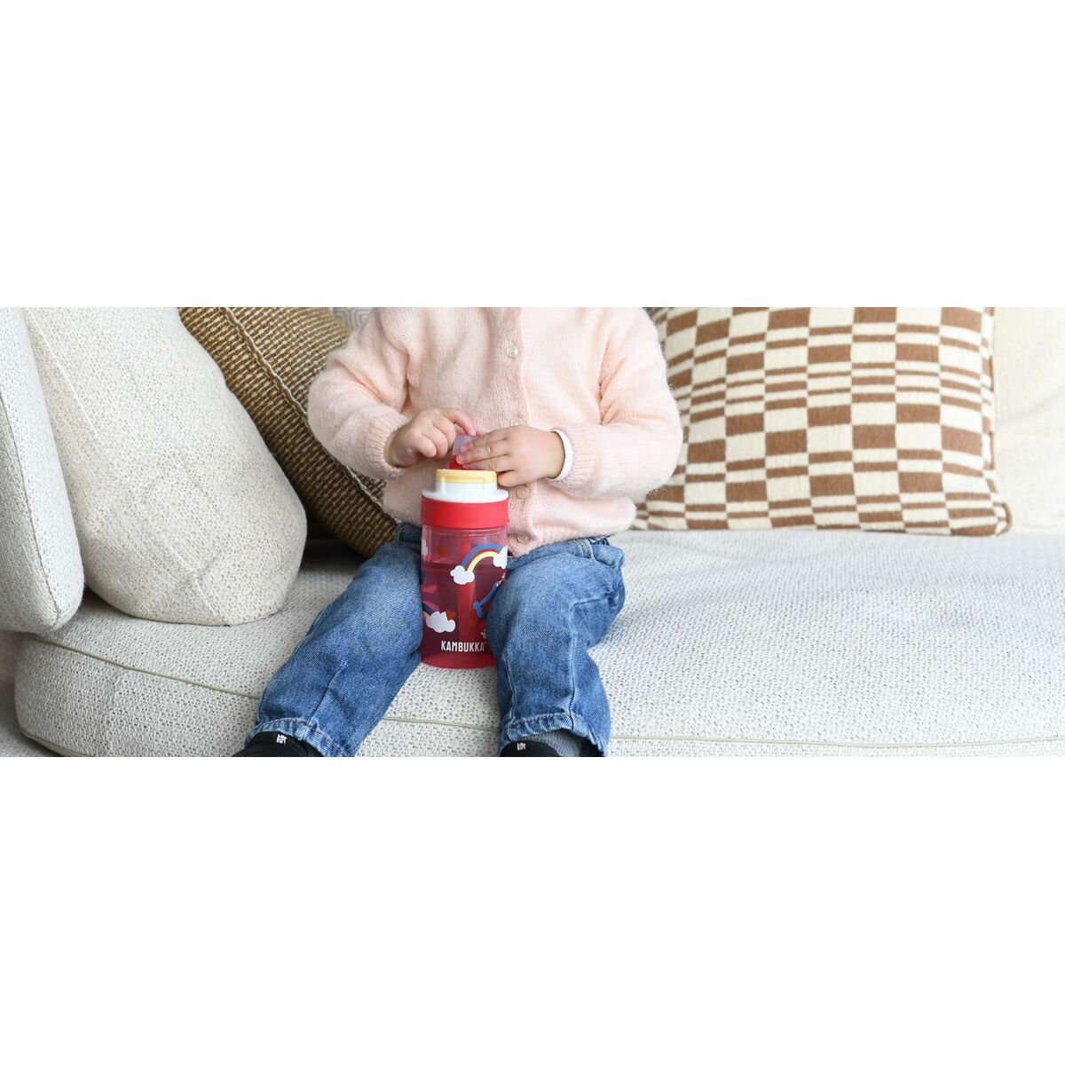 Ein Kind in einem rosa Pullover und Jeans sitzt und hält eine rote Wasserflasche mit Regenbogenmuster auf einer weißen Couch mit Kissen.