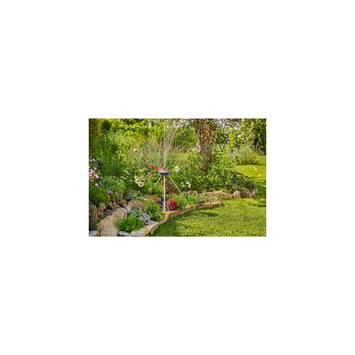 Ein Gartensprenger auf einem Dreibein bewässert ein Blumenbeet und Rasen. Im Hintergrund sind Bäume und blühende Blumen.