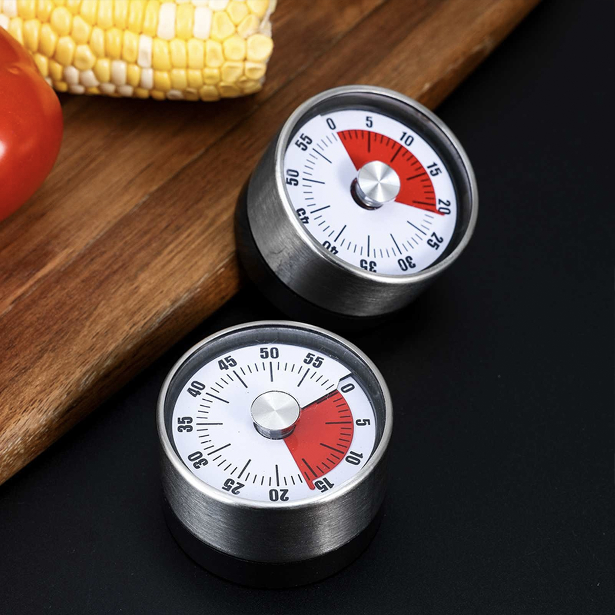 Zwei silberne Küchenwecker mit roten Abschnitten, neben Mais und einer Tomate auf einem Holzbrett, vor schwarzem Hintergrund.