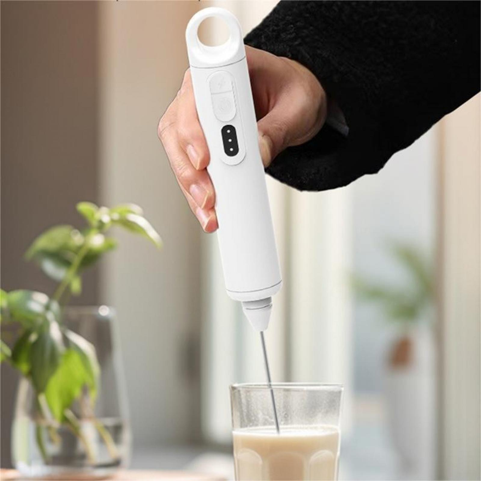 Eine Hand hält einen weißen Aufschäumer über einem Glas Flüssigkeit. Im Hintergrund sind eine Pflanze und ein Fenster zu sehen.
