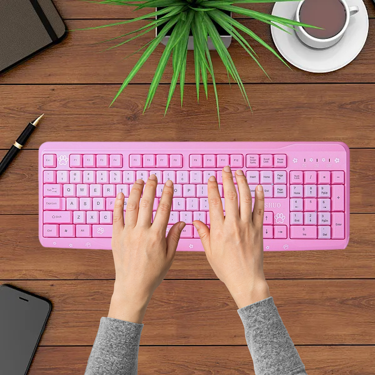 Hände tippen auf einer rosa Tastatur. Eine Tasse Kaffee, eine Pflanze und ein Telefon liegen ebenfalls auf dem Holztisch.