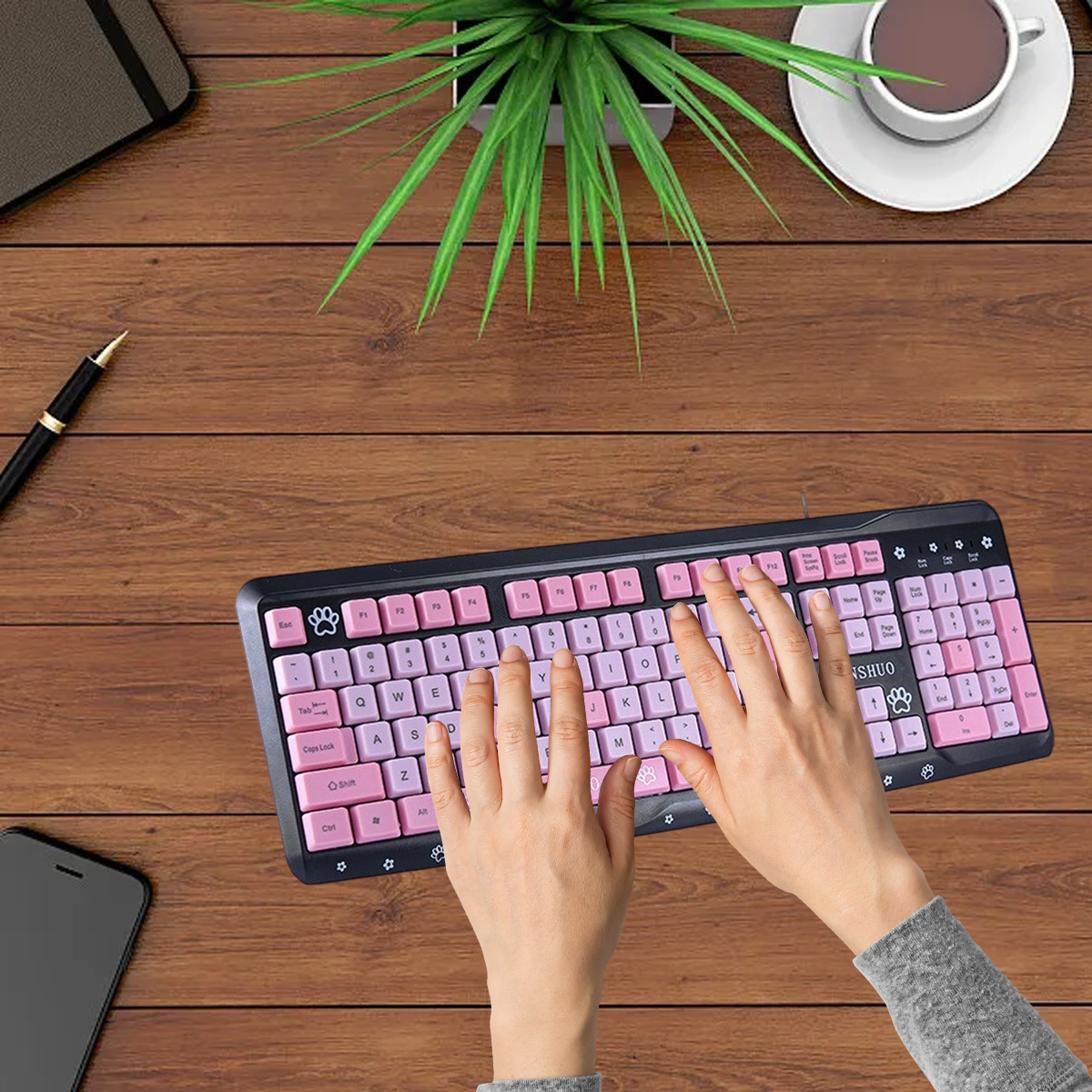 Ein Schreibtisch-Setup mit einer rosa Tastatur, einer Tasse Kaffee, einer Pflanze, einem Stift, einem Telefon und Händen, die auf der Tastatur tippen.