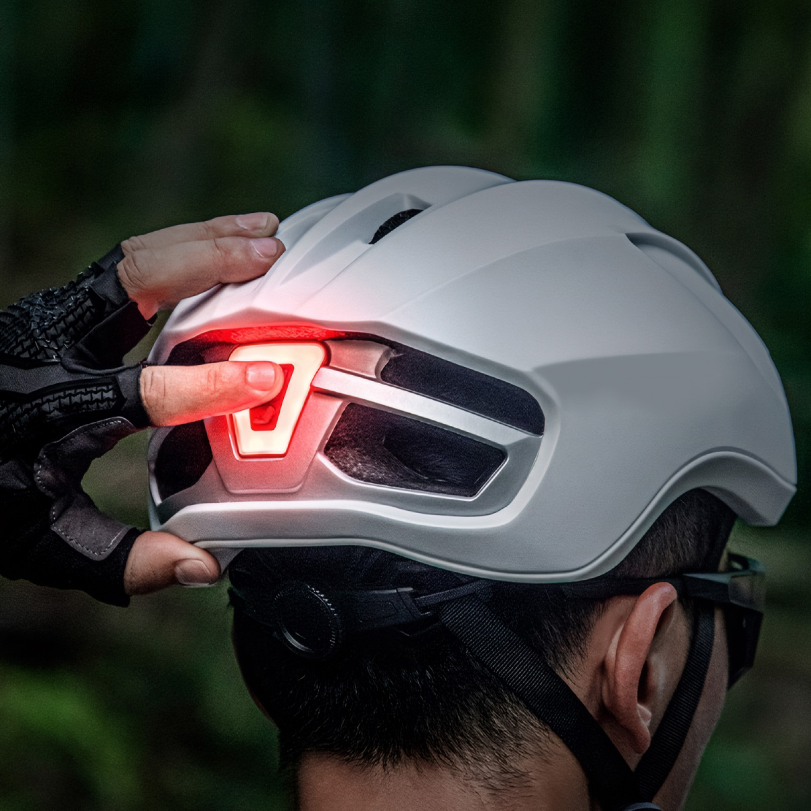 Die Hand einer Person aktiviert das rote Licht auf der Rückseite eines weißen Fahrradhelms. Grüner verschwommener Hintergrund.