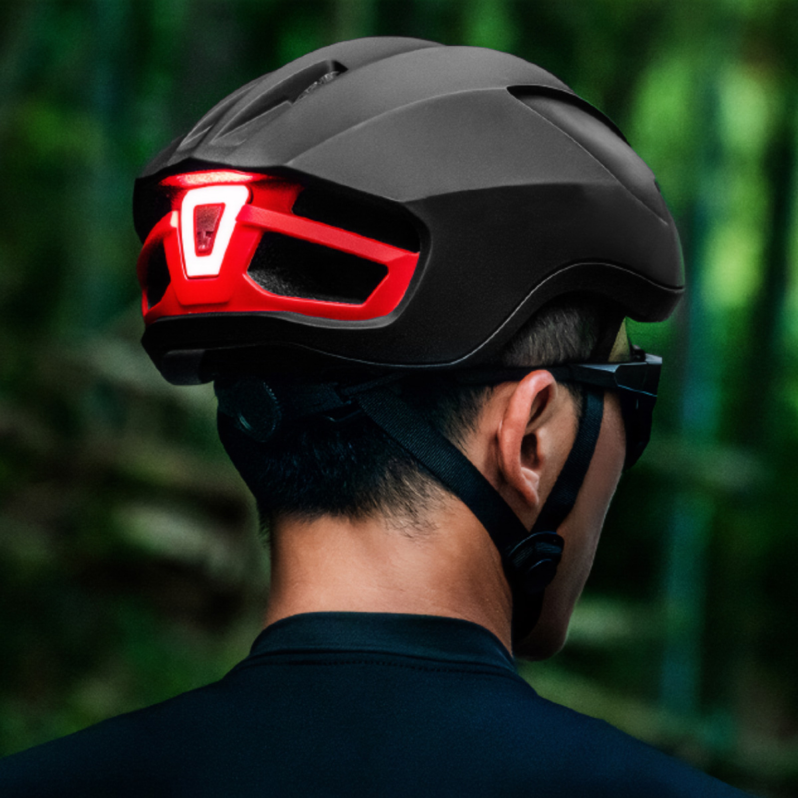 Eine Person trägt einen schwarzen Fahrradhelm mit rotem Licht hinten. Der Hintergrund ist verschwommenes grünes Laub.