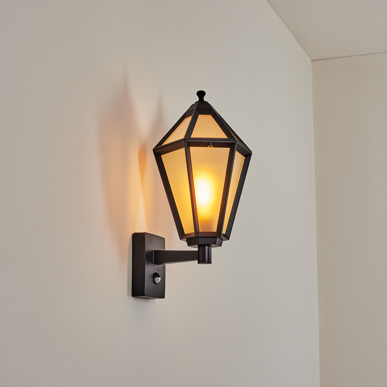Eine schwarze Wandleuchte mit geometrischem Design, von innen beleuchtet. Sie ist an einer hellen Wand befestigt.