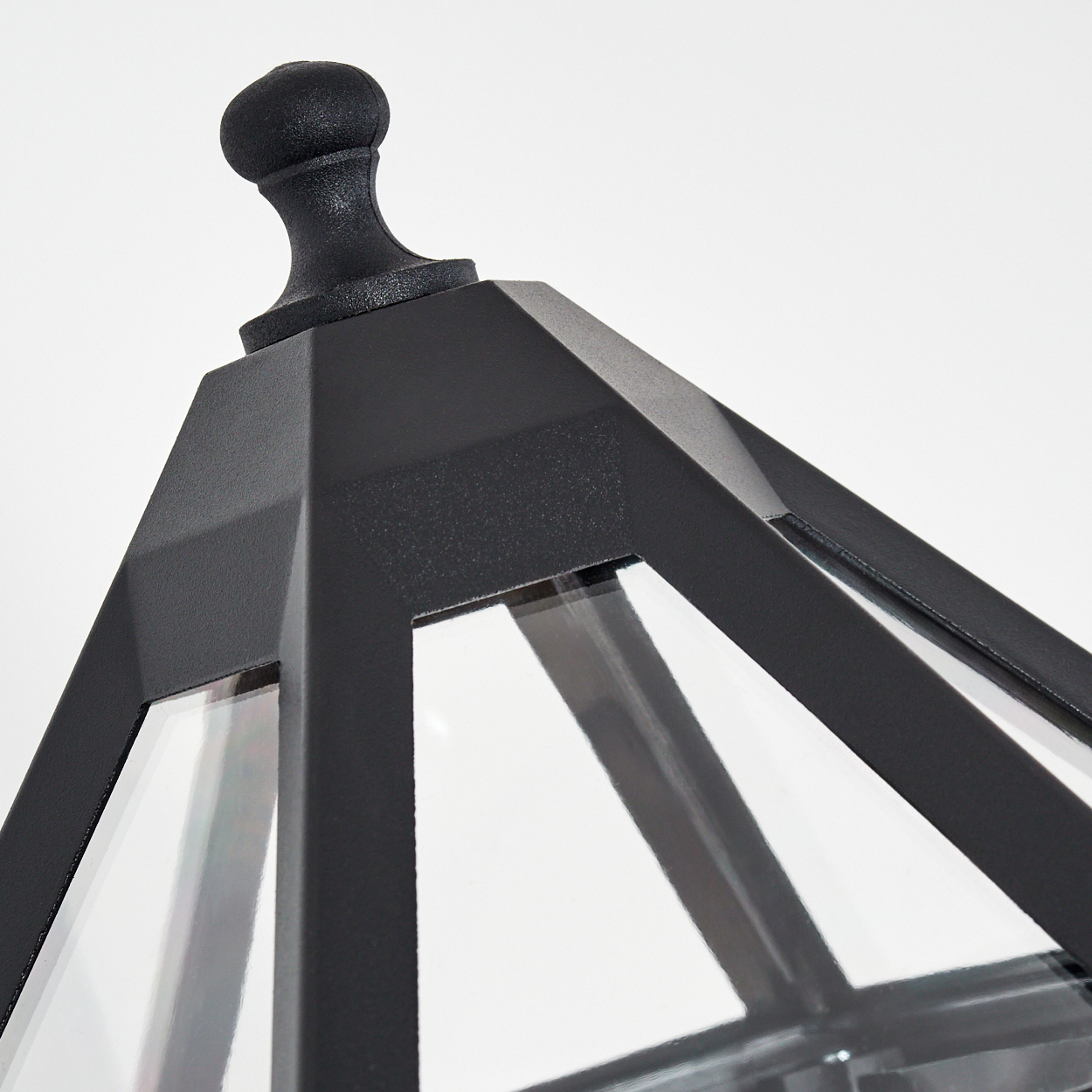 Nahaufnahme einer schwarzen, geometrischen Laternenoberseite. Sie hat eine dekorative Spitze, Glasscheiben und einen schwarzen Rahmen vor weißem Hintergrund.