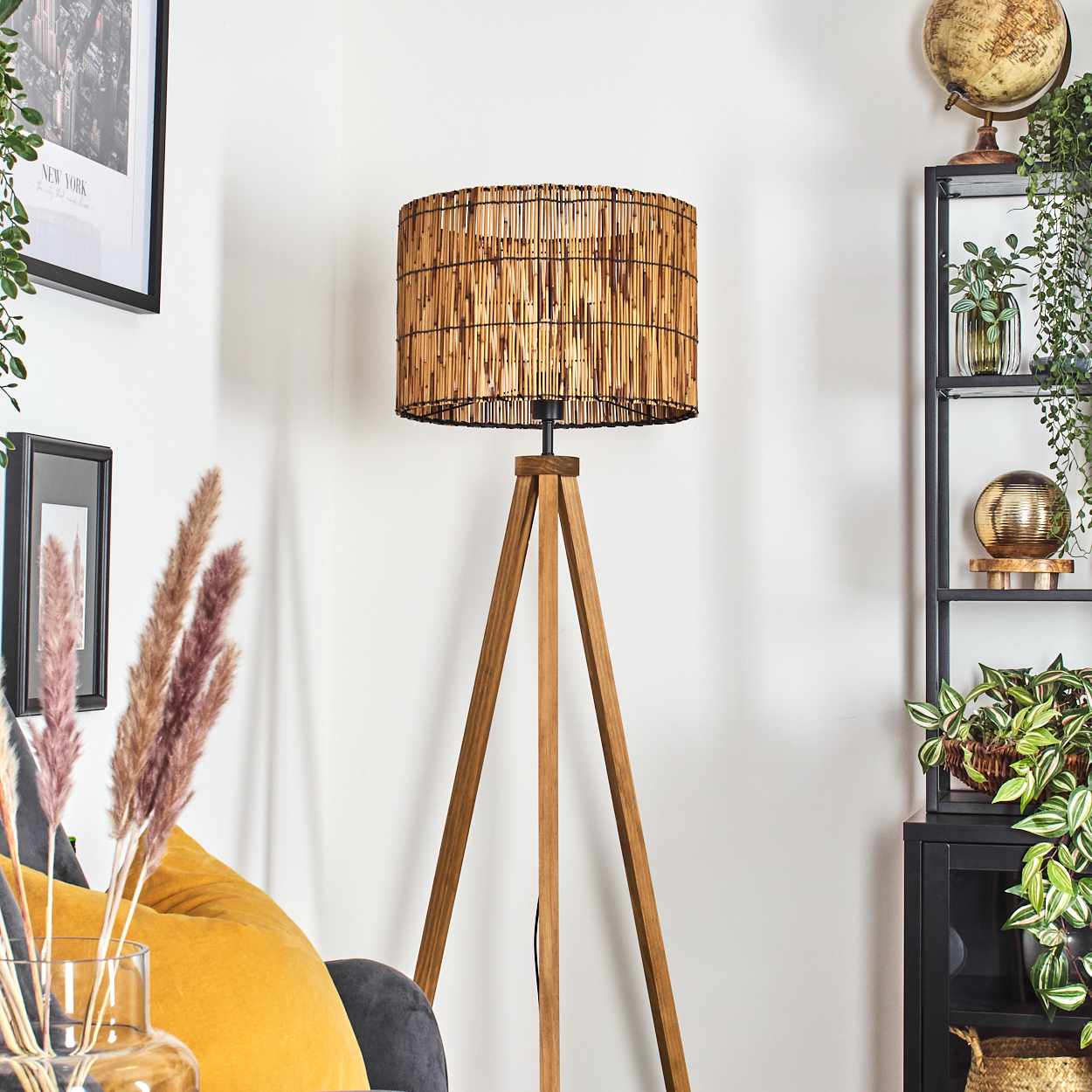 Eine Stehlampe mit Holzstativ und gewebtem Lampenschirm steht in einem Raum. Dahinter ein Regal mit Pflanzen und Globus. Ein gelbes Kissen ist sichtbar.
