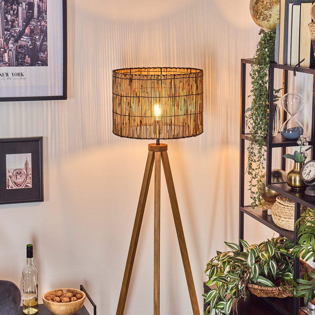 Eine Stehlampe mit gewebtem Schirm steht vor einem Regal mit Dekoration und Pflanzen. Ein Bild von NYC und eine Schüssel mit Nüssen sind sichtbar.