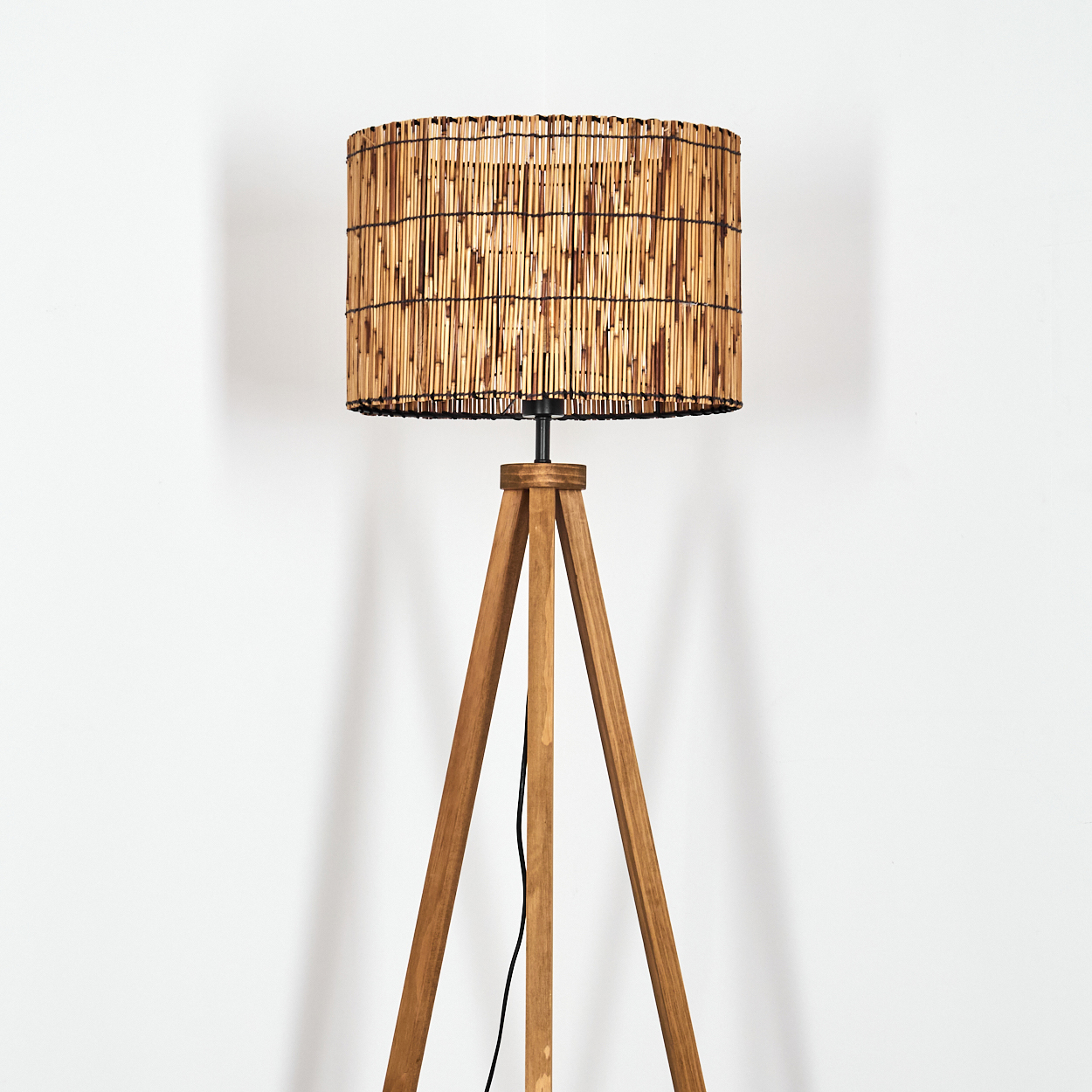 Eine Stehlampe mit Holzstativ und Bambus-Lampenschirm steht vor einer weißen Wand. Ein schwarzes Kabel ist sichtbar.