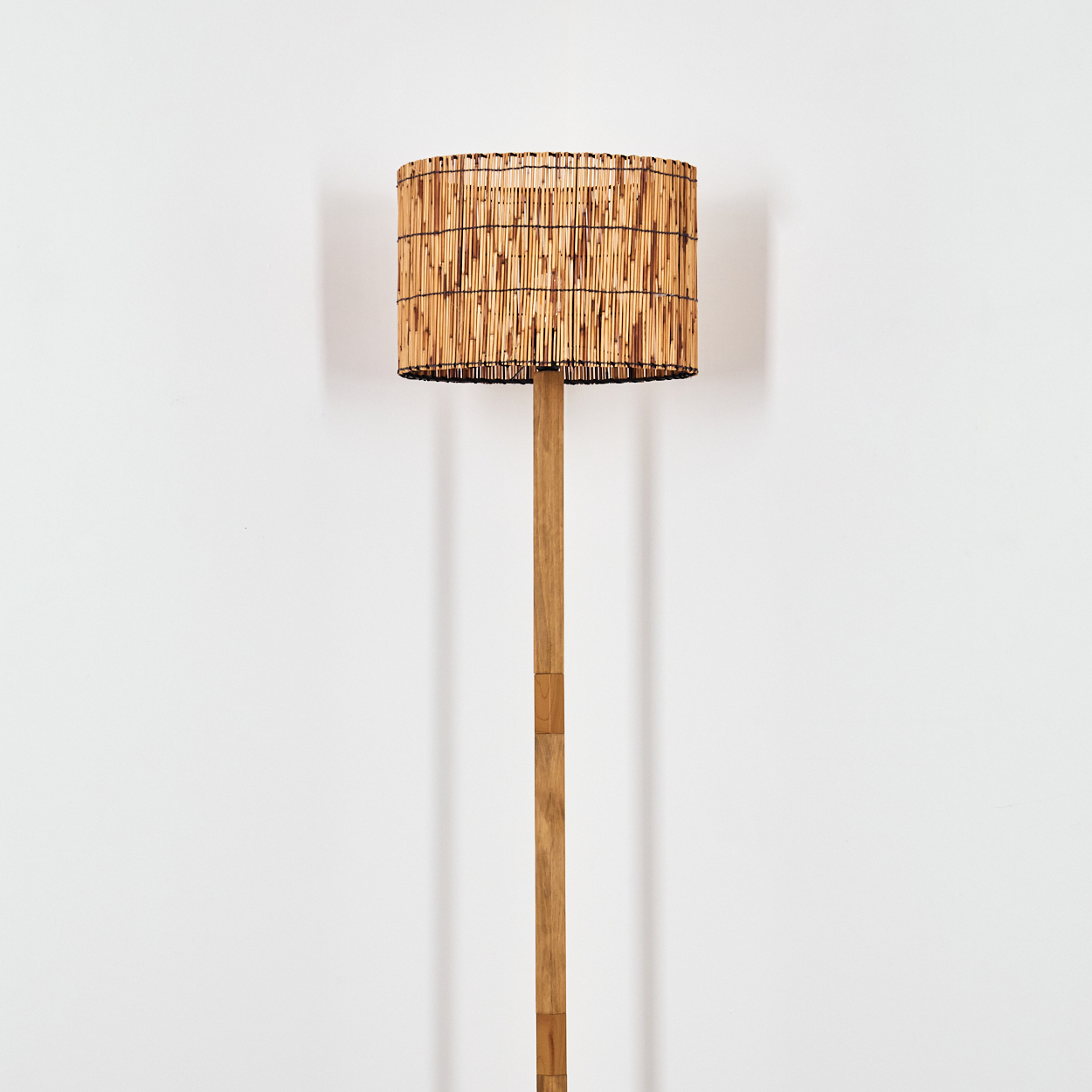 Ein Bambus-Lampenschirm sitzt auf einem Holzpfahl. Der Schirm ist rechteckig, und der Hintergrund ist eine schlichte weiße Wand.