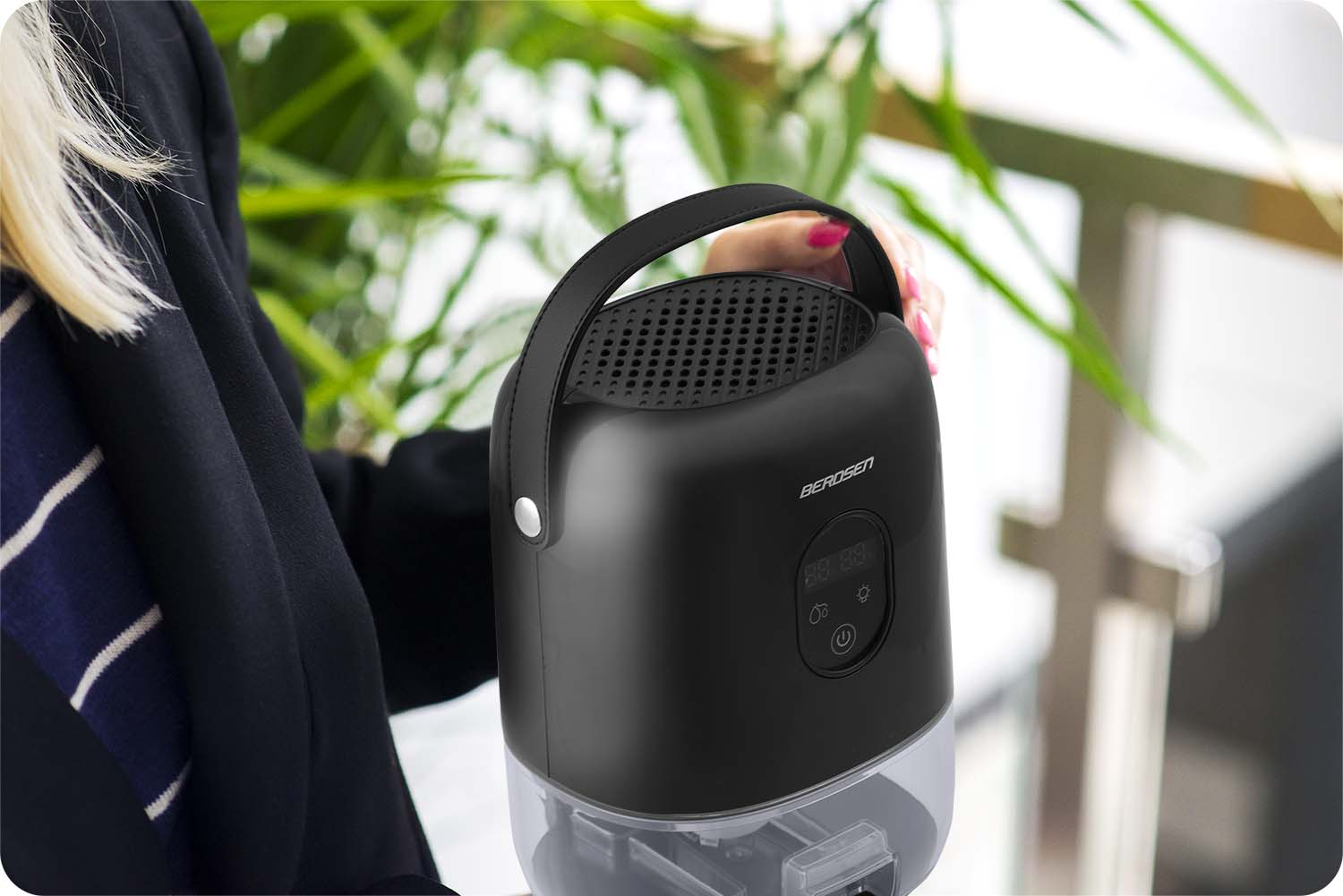 Osoba trzyma czarny oczyszczacz powietrza Berosen. Oczyszczacz ma uchwyt i wyświetlacz. W tle zielone liście.