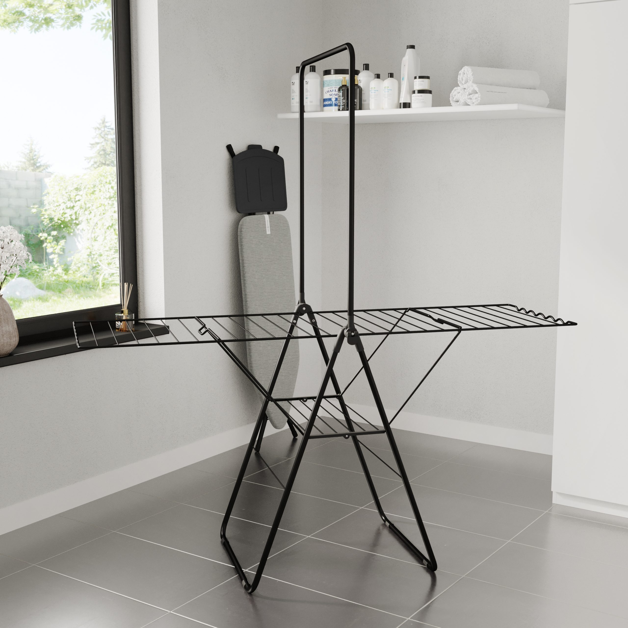 A black clothes drying rack in a room, next to a window. An ironing board is also visible, along with laundry supplies.