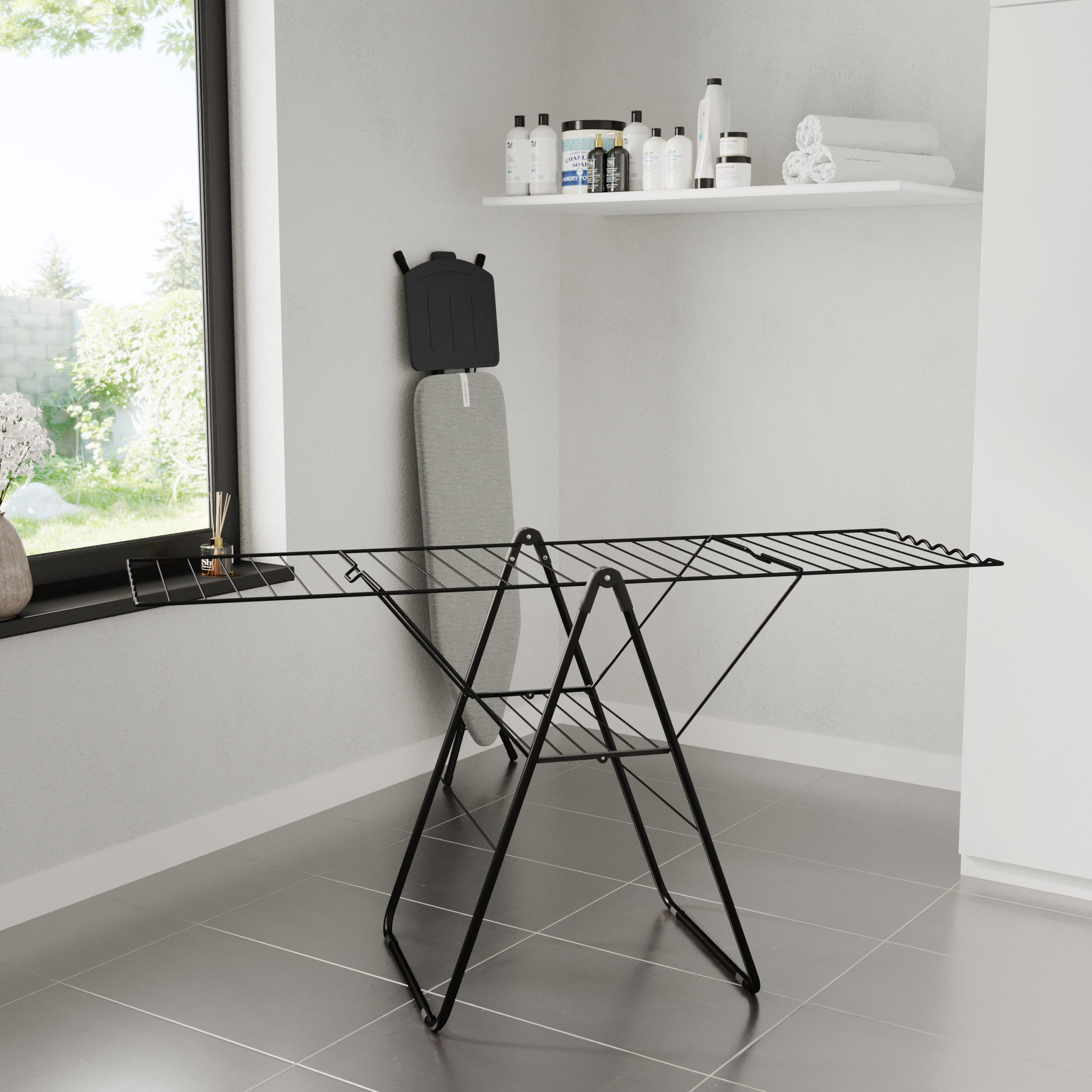 A room with a black clothes drying rack, ironing board, and a window looking out to greenery.
