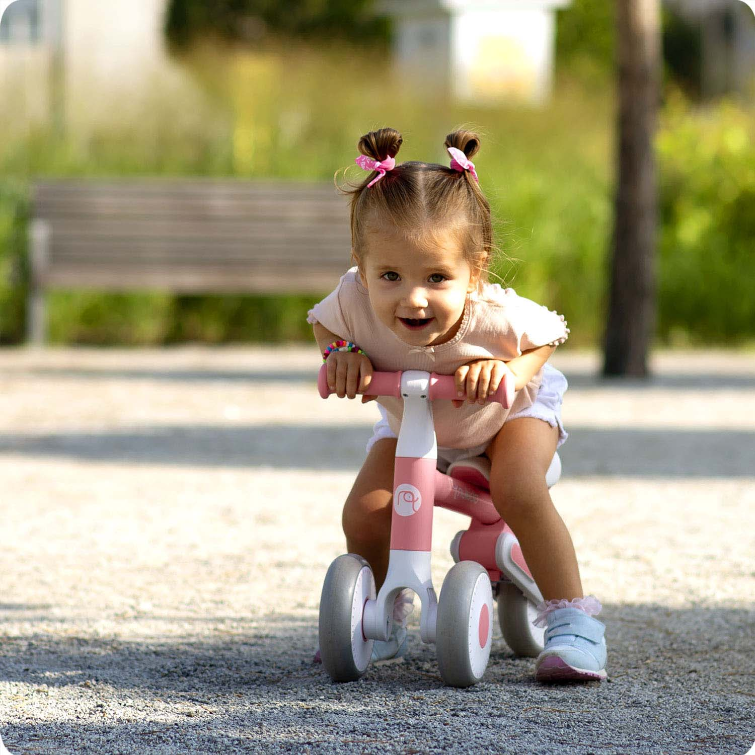 Młoda dziewczyna z kucykami jeździ na różowo-białym rowerku biegowym po żwirowej ścieżce, uśmiechając się. Tło obejmuje ławkę i drzewa.