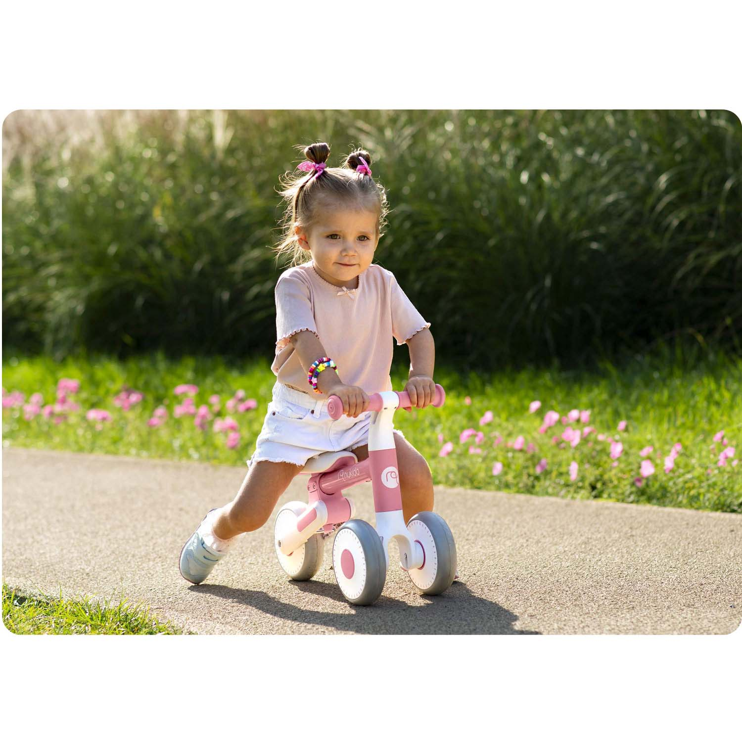 Mała dziewczynka jeździ na różowo-białym rowerku biegowym po utwardzonej ścieżce. Ma na sobie różową koszulkę, białe szorty i ma koki.