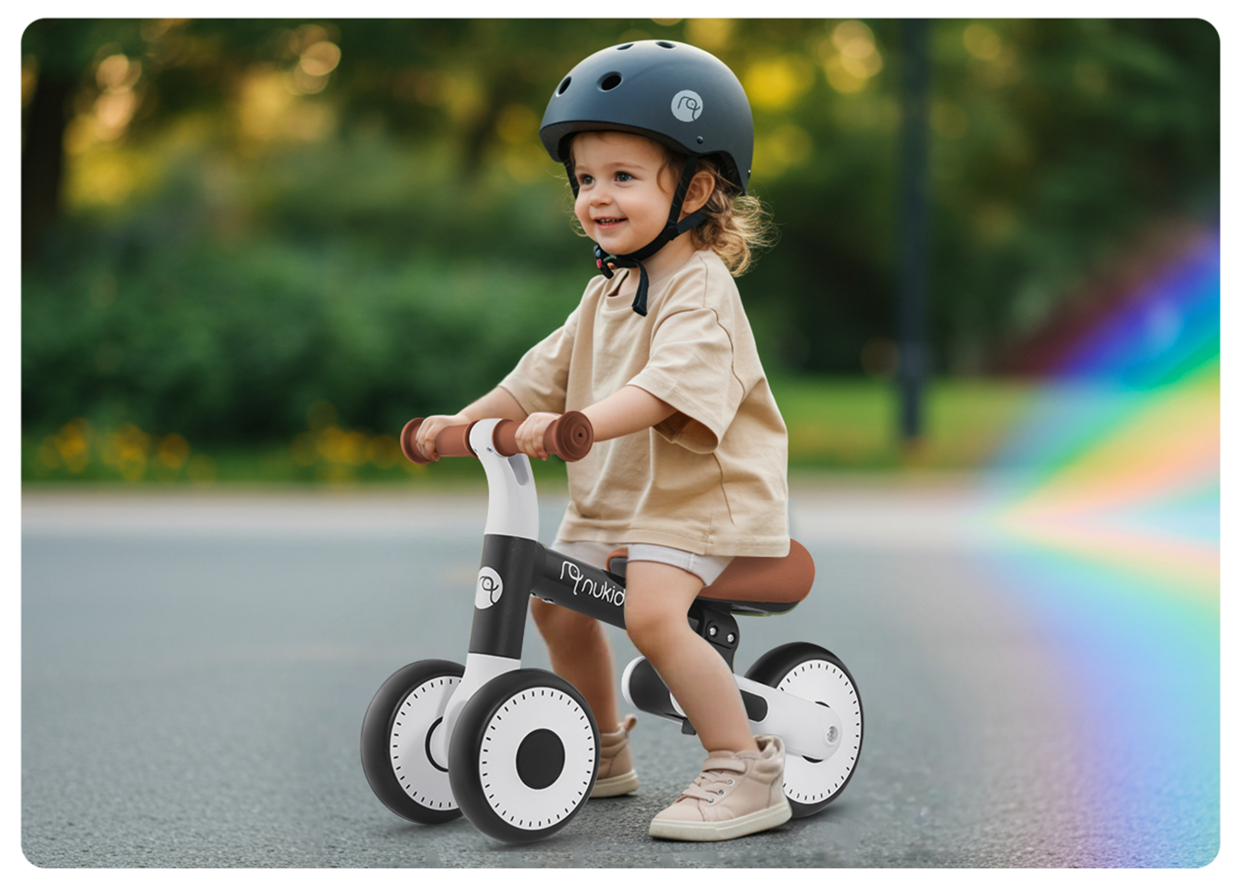 Dziecko w kasku jeździ na rowerze biegowym po drodze. Zielone drzewa w tle.