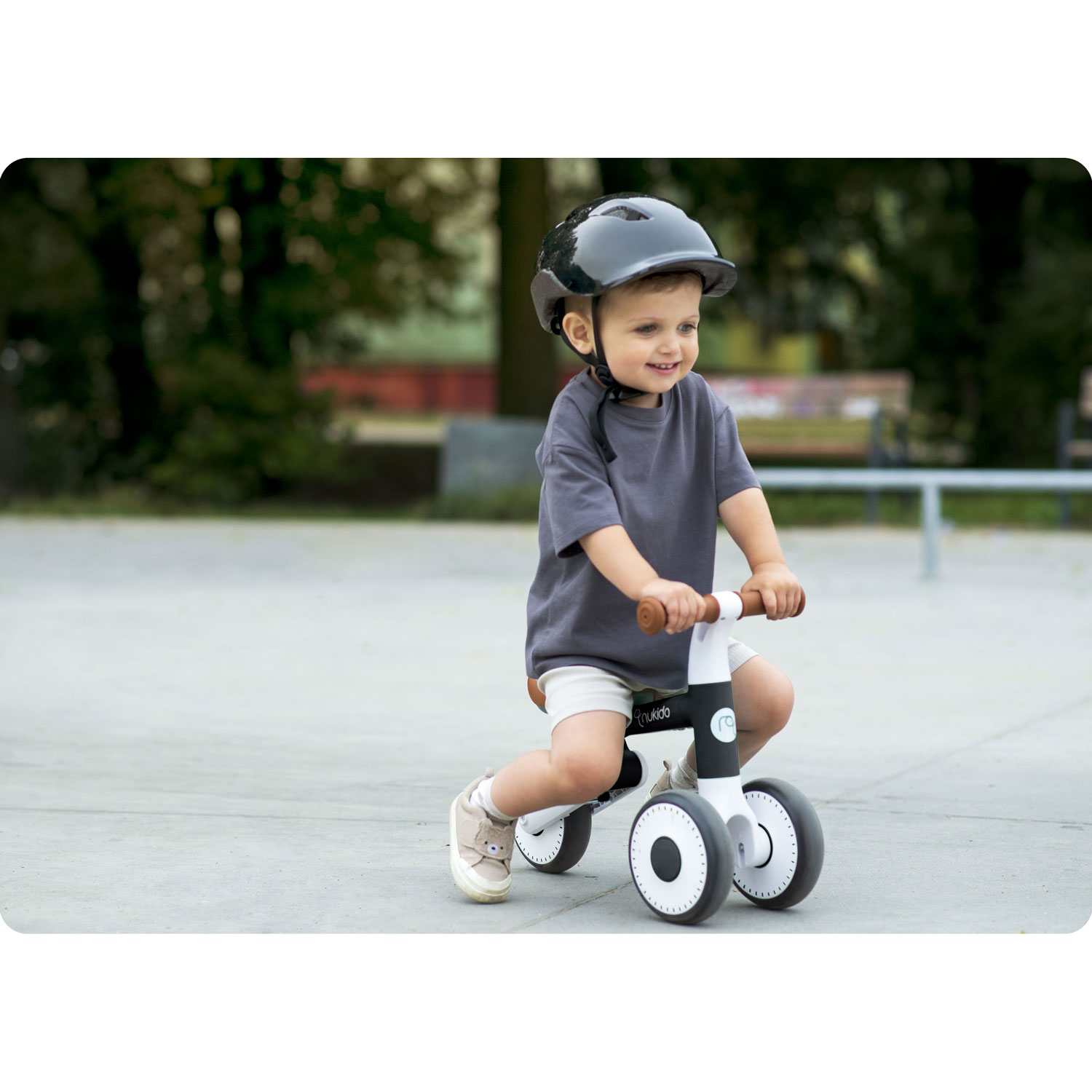 Dziecko w kasku jeździ na rowerku biegowym po betonowej nawierzchni. Drzewa i ławka w rozmytym tle.