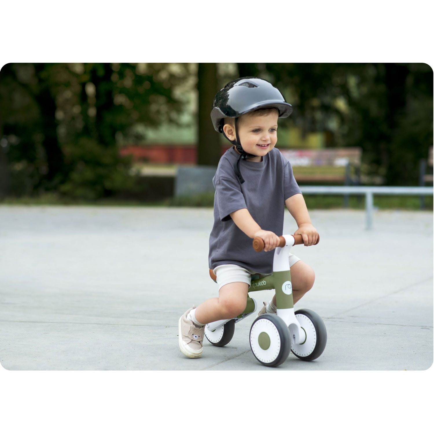 Małe dziecko w kasku jeździ na rowerze biegowym. Uśmiecha się na tle parku, drzew i ławki.