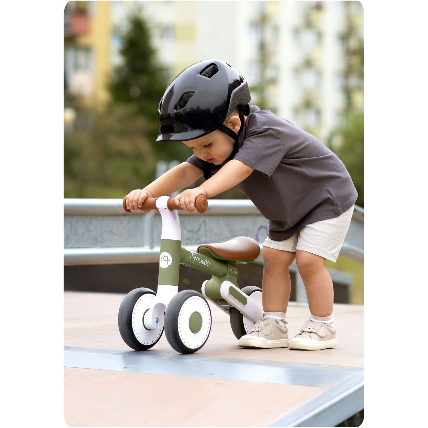 Dziecko w czarnym kasku pcha zielono-biały rowerek biegowy. Dziecko ma na sobie szarą koszulkę i białe spodenki.