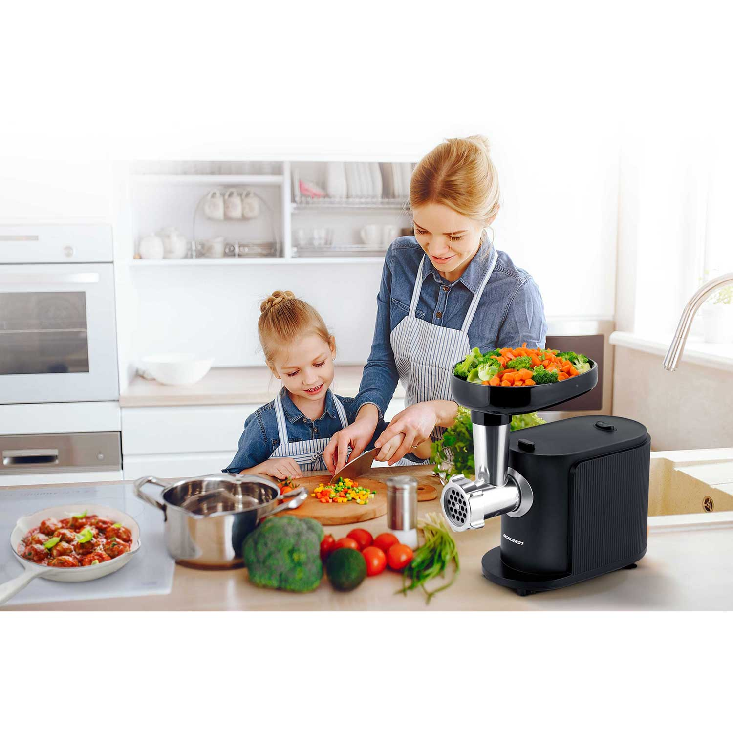 Kobieta i dziecko kroją warzywa. Po prawej stronie maszynka do mięsa z warzywami. Po lewej patelnia z klopsikami.