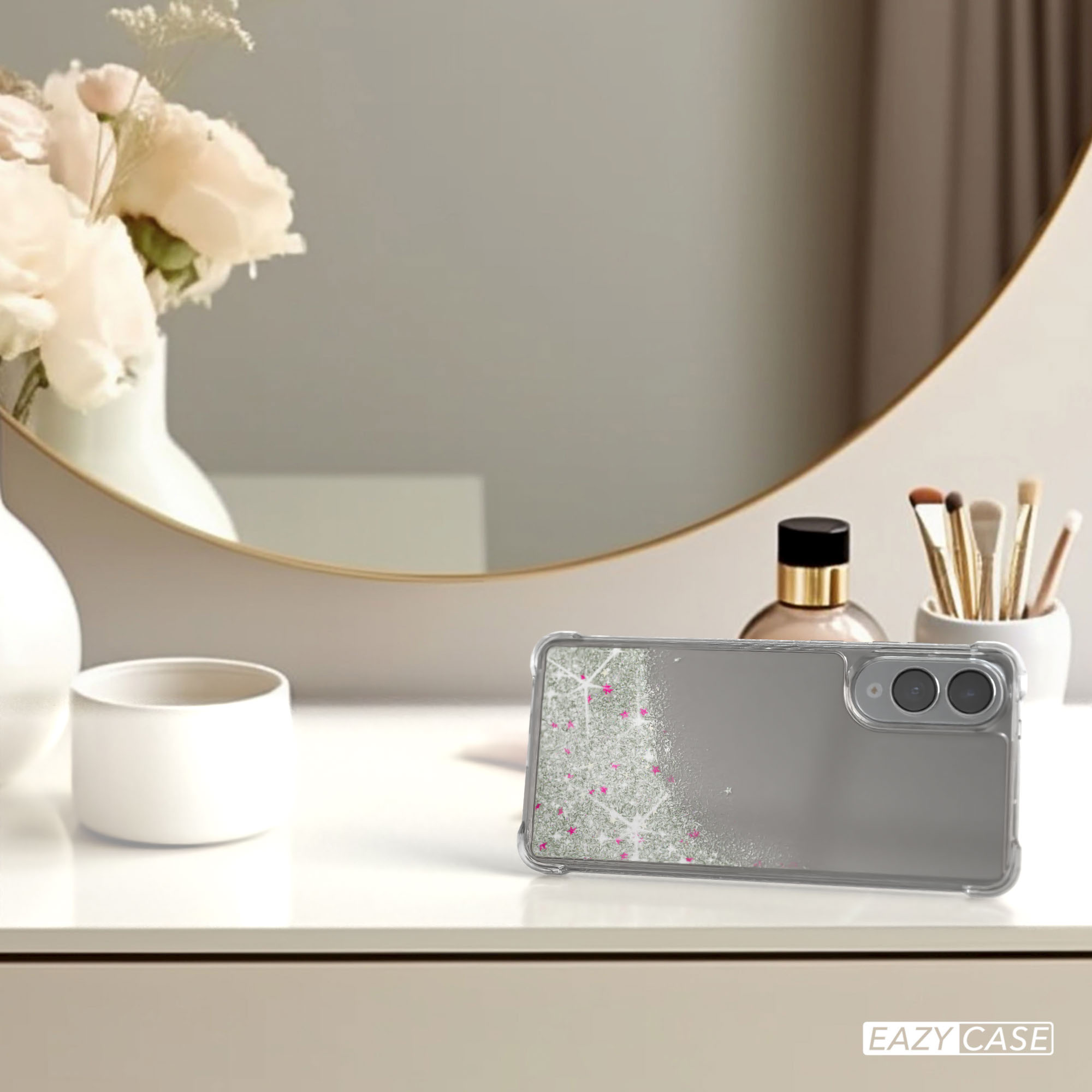 Eine Handyhülle mit Design, ein Spiegel, Blumen und Make-up-Pinsel auf einer weißen Oberfläche. Der Hintergrund ist verschwommen.