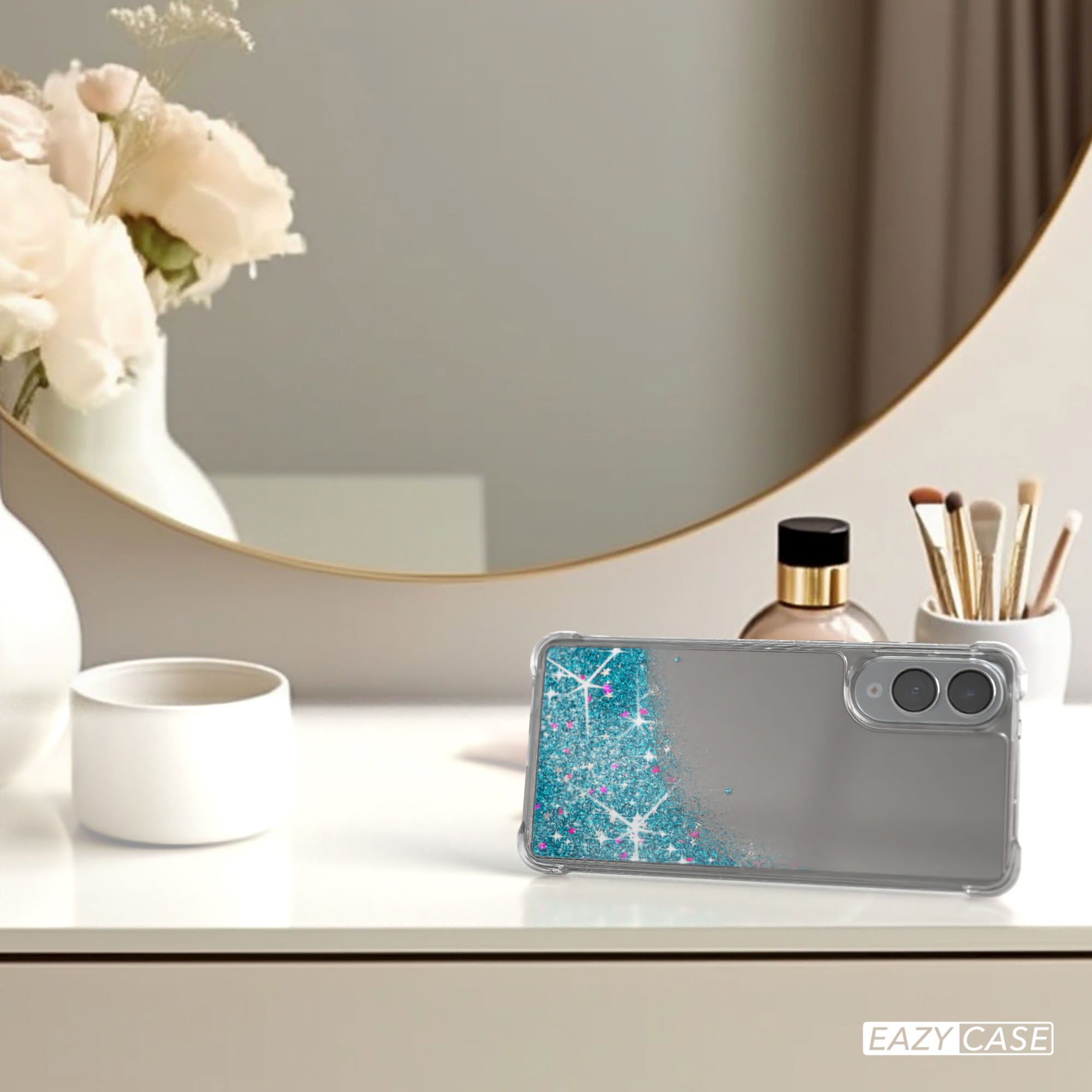 Eine Handyhülle mit blauem und rosa Glitzerdesign liegt auf einer weißen Oberfläche. Hintergrund: Spiegel, Blumen und Make-up.
