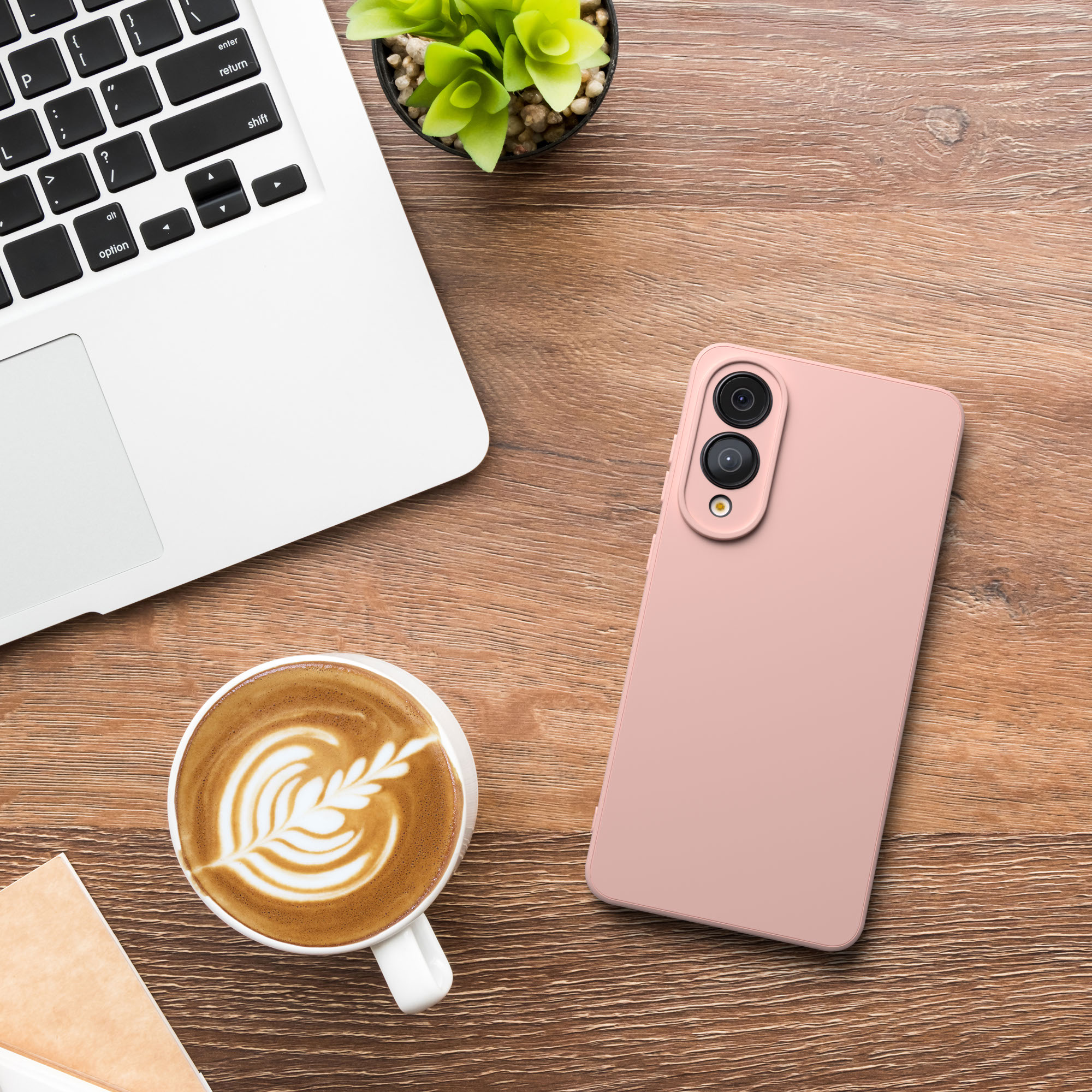 Ein Laptop, Kaffee mit Latte Art, ein rosa Telefon und eine kleine Pflanze auf einem Holztisch.