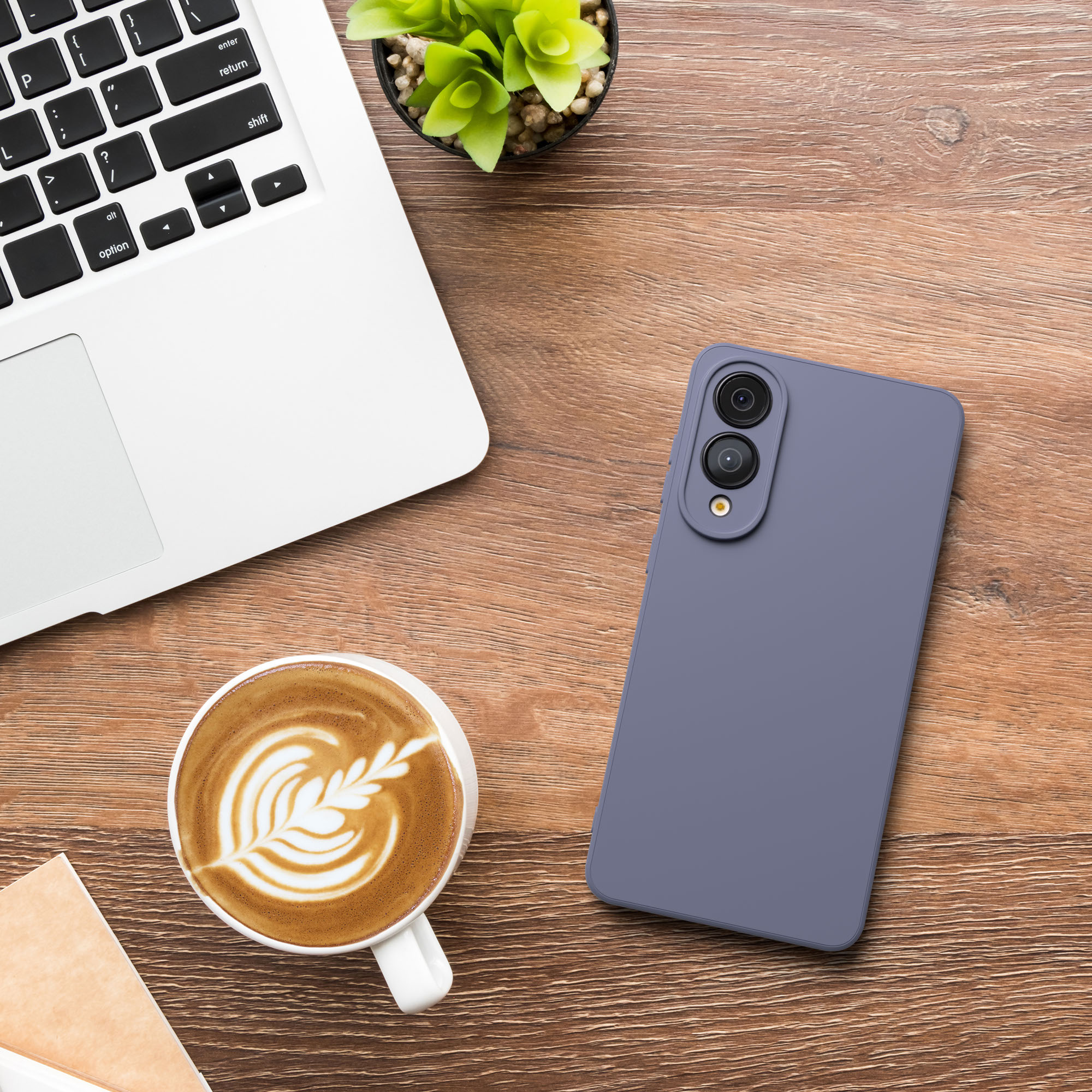 Auf einem Holztisch liegen ein Laptop, ein Telefon und eine Kaffeetasse. Eine Pflanze ist ebenfalls vorhanden.