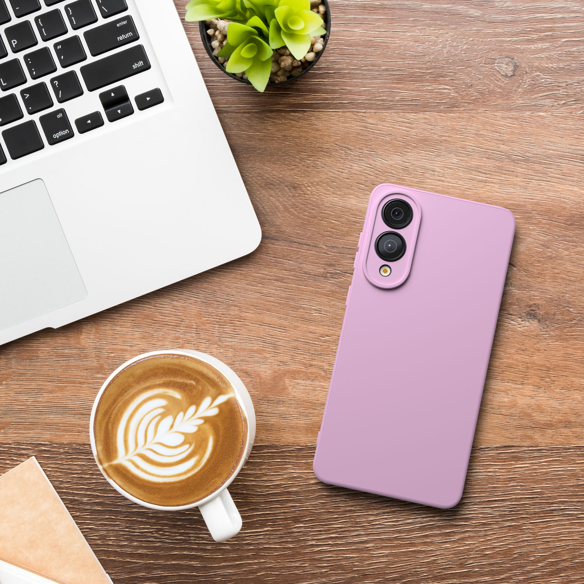 Ein Laptop, ein pinkes Telefon, eine Kaffeetasse und eine Pflanze liegen auf einem Holzschreibtisch. Der Kaffee hat Latte Art.